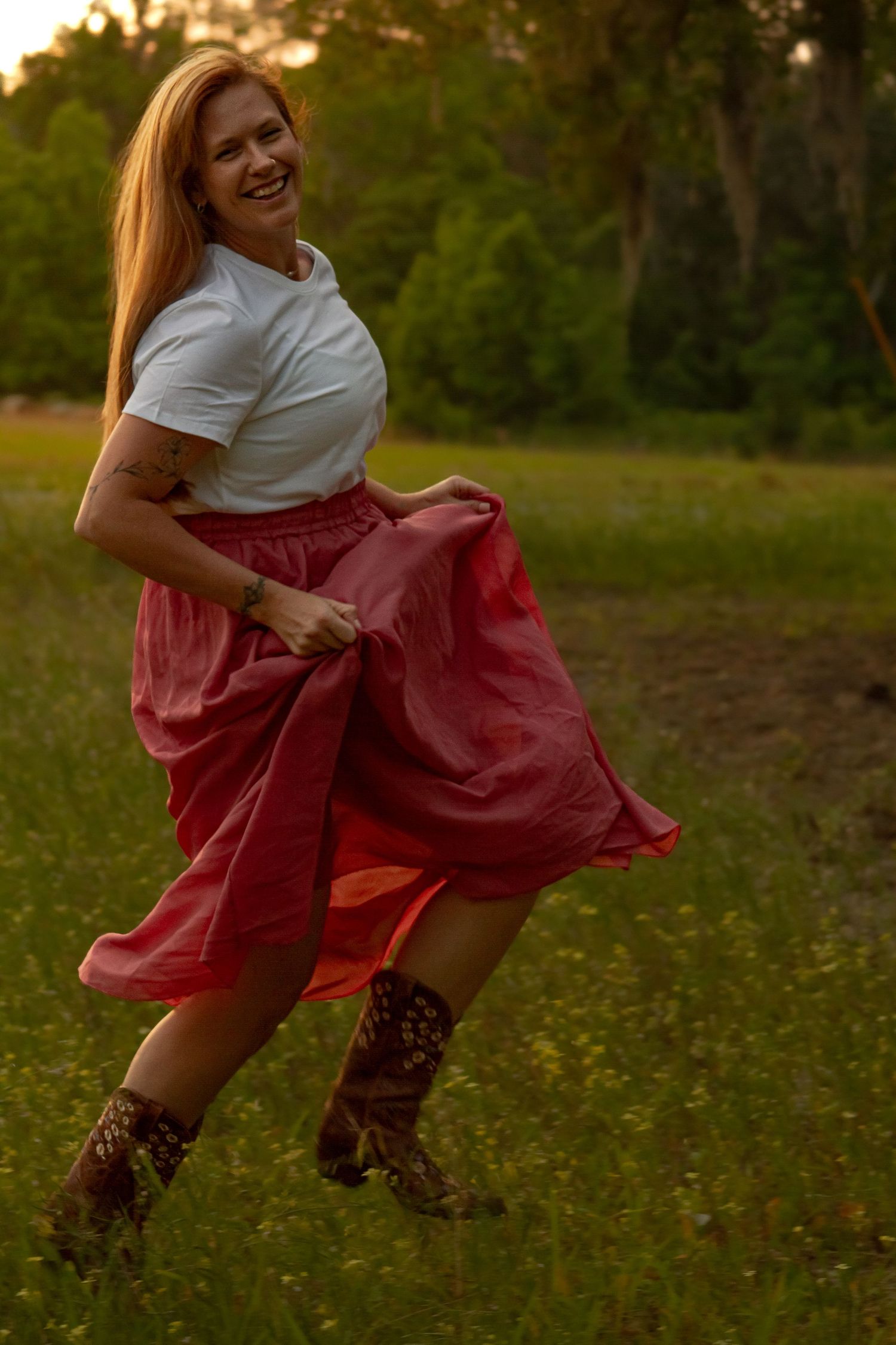 Laughing redhead woman in white top and red skirt twirling in a green meadow at golden hour.
