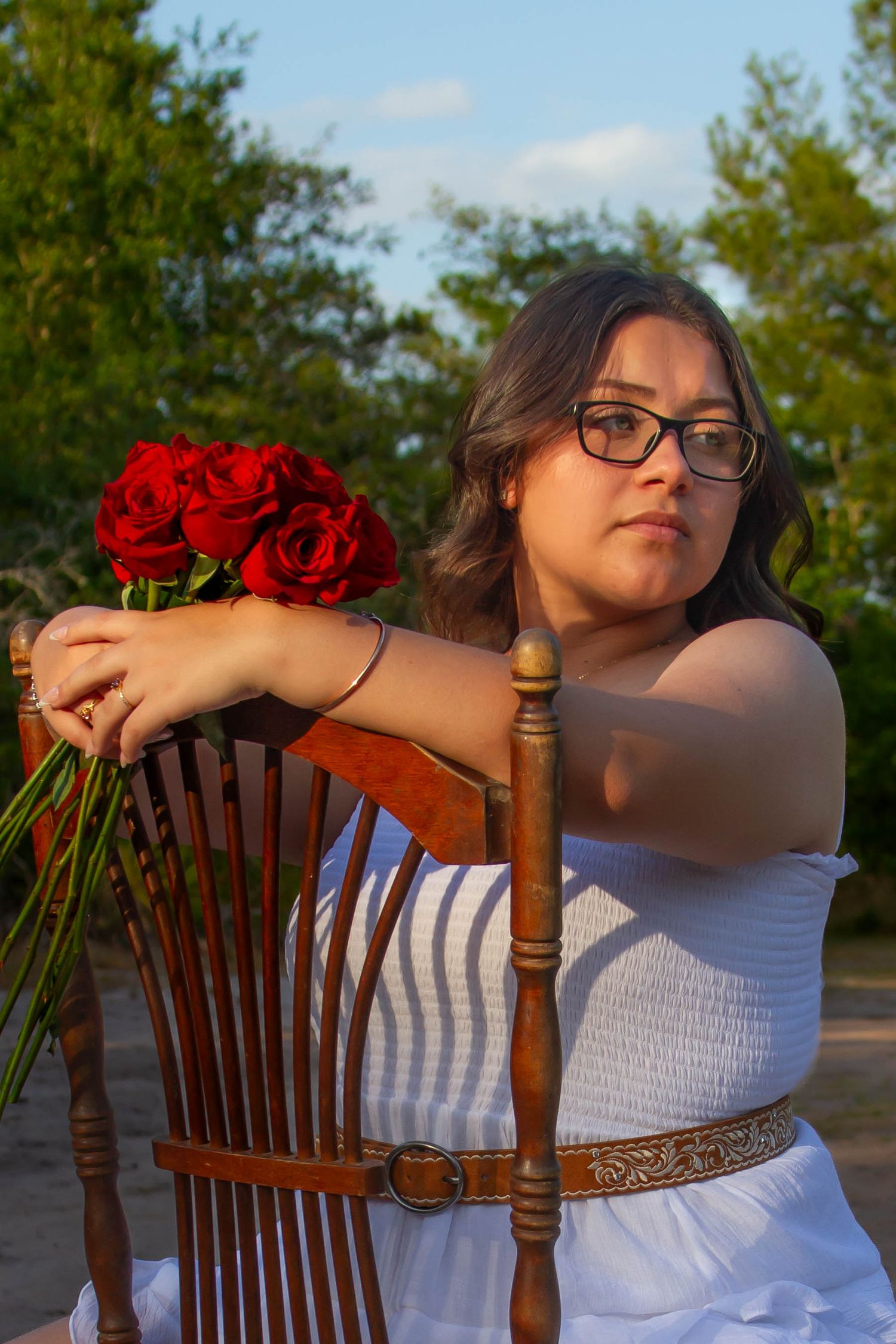 Young woman with glasses holding red roses, leaning on wooden chair in golden hour outdoor light.