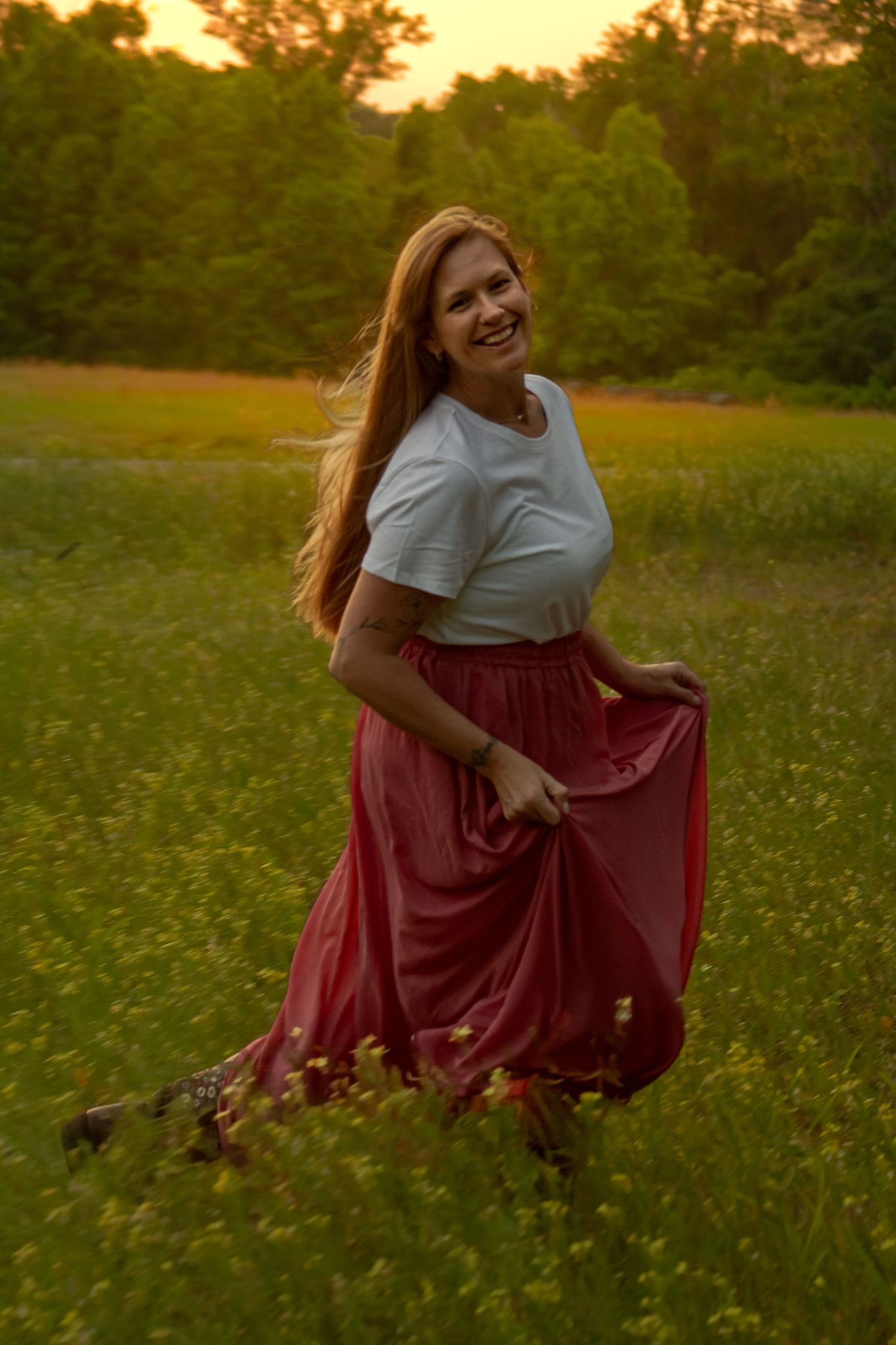 Smiling redhead woman in white tee and flowing red maxi skirt twirling in a sunny wildflower meadow.