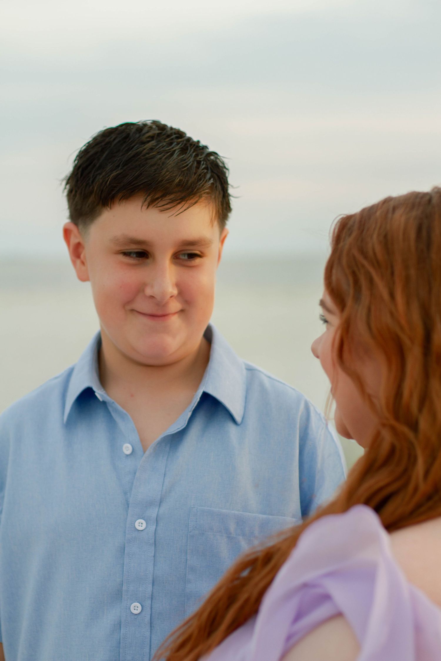 A smiling teenage boy in a light blue shirt gazes at a red-haired mother near the ocean.