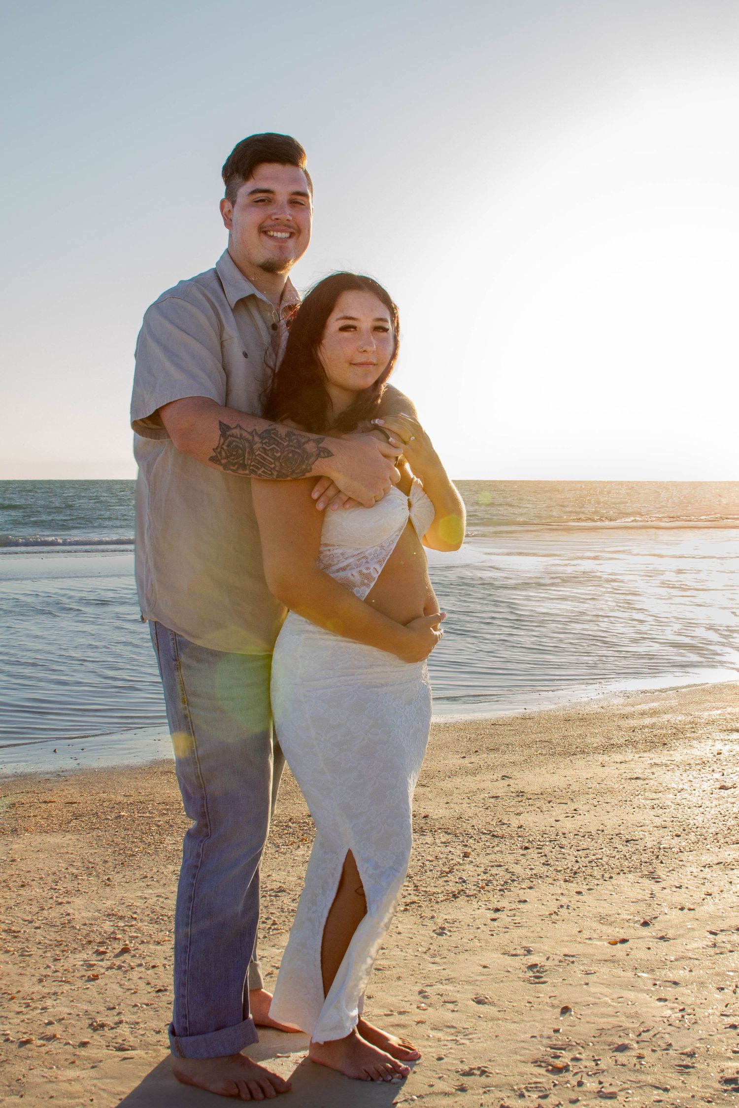 Couple embracing on beach at sunset, woman in white dress showing baby bump.