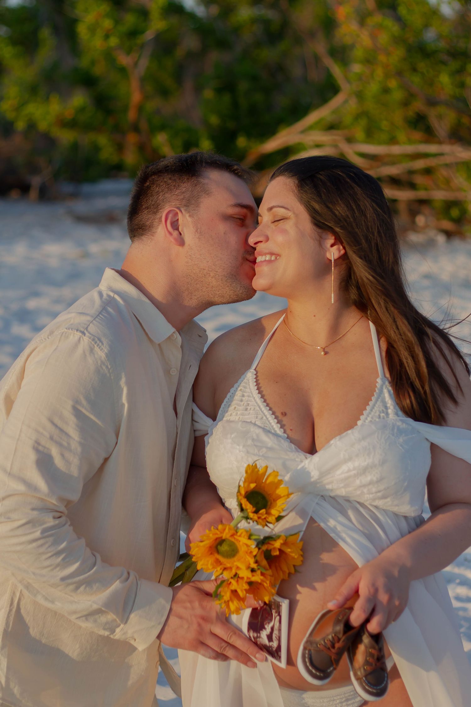 Man kisses smiling pregnant woman's cheek as she holds sunflowers and ultrasound photos on sunny beach.