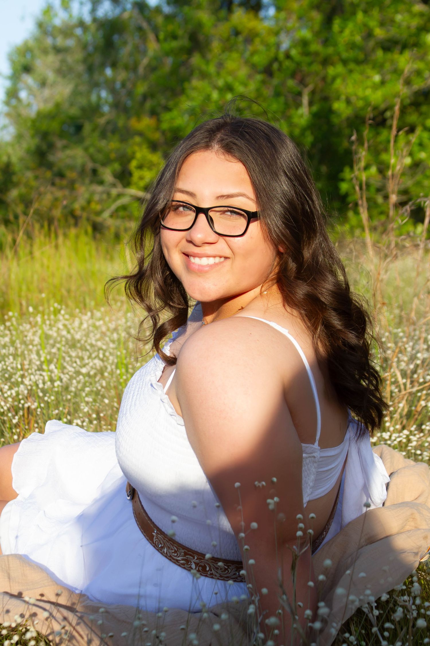 Smiling young woman in white dress sitting in wildflower field in golden sunlight.
