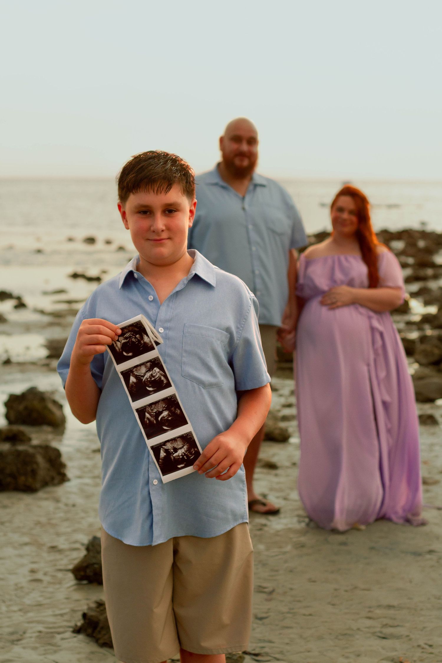 Young boy holds ultrasound strip on beach with pregnant mother and father posing behind him.