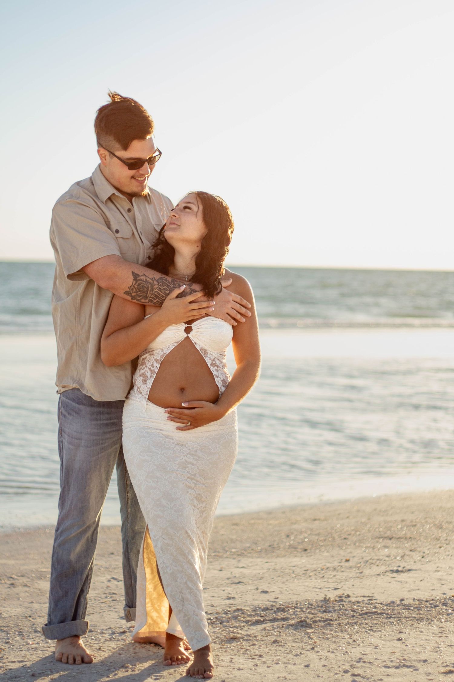 Couple sharing a tender embrace on the beach at sunset, pregnant woman in white lace skirt smiling up.