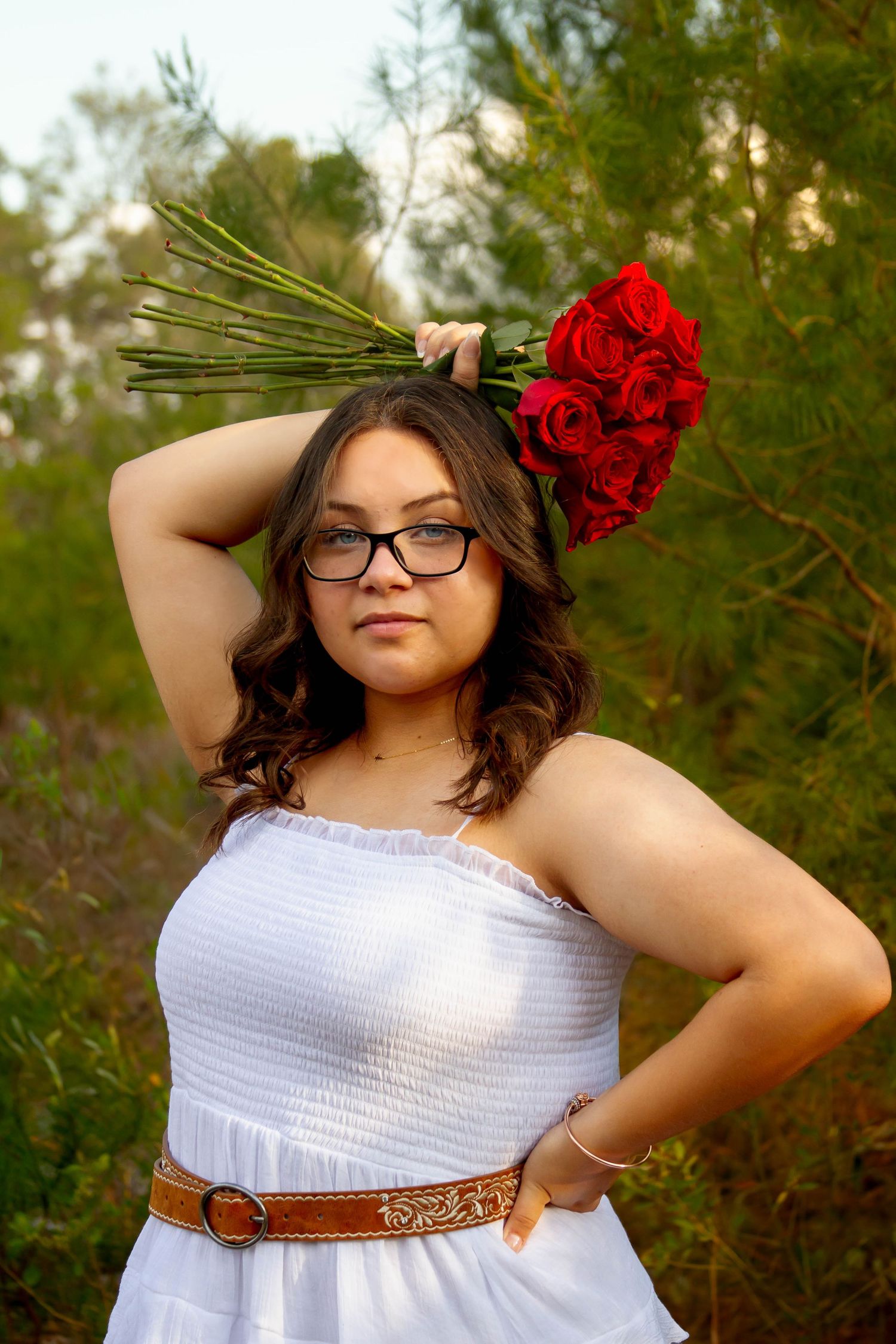 Young woman in white dress holding red roses bouquet over shoulder in nature setting.