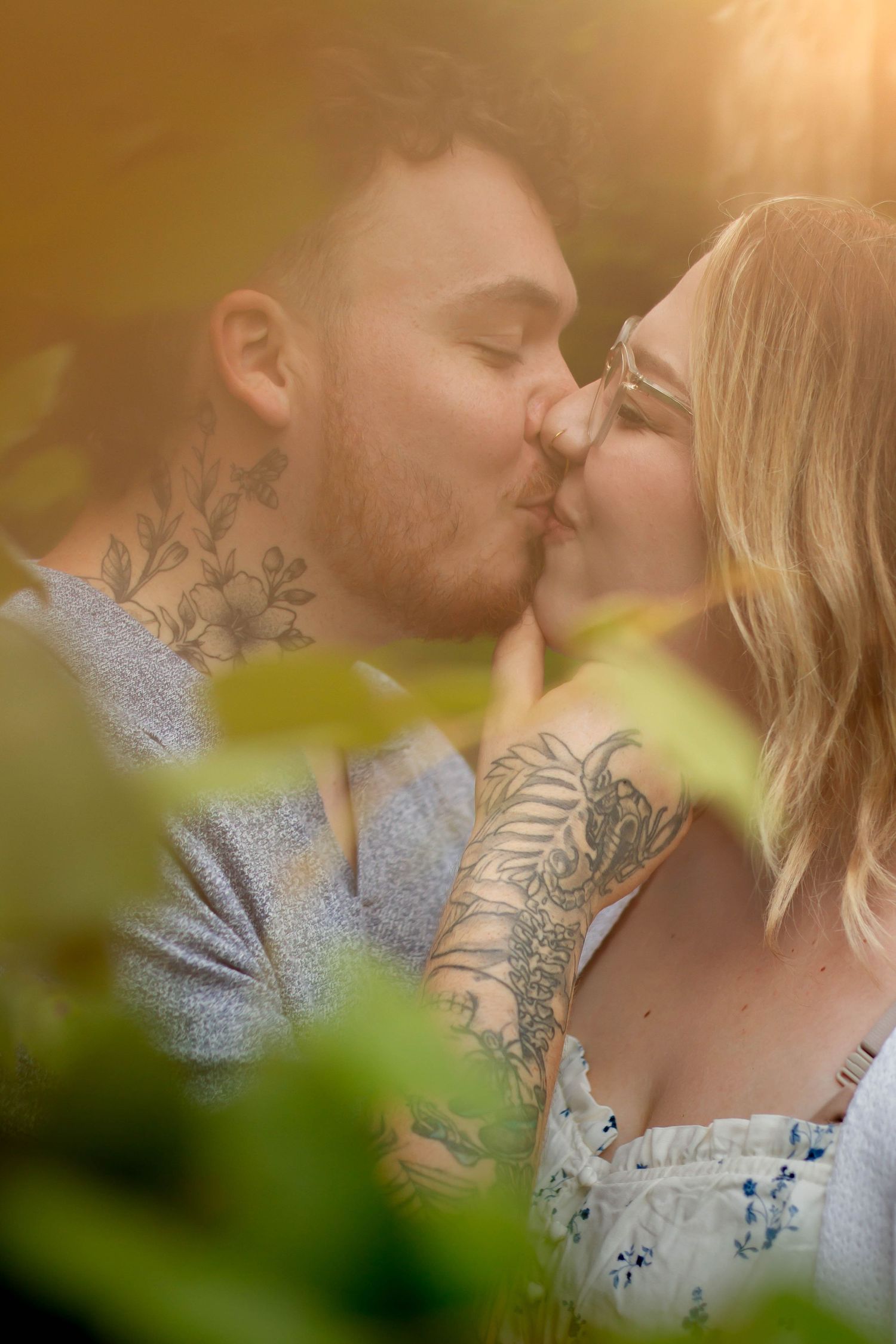 A tattooed couple shares a romantic kiss outdoors, surrounded by lush green leaves in warm golden sunlight.