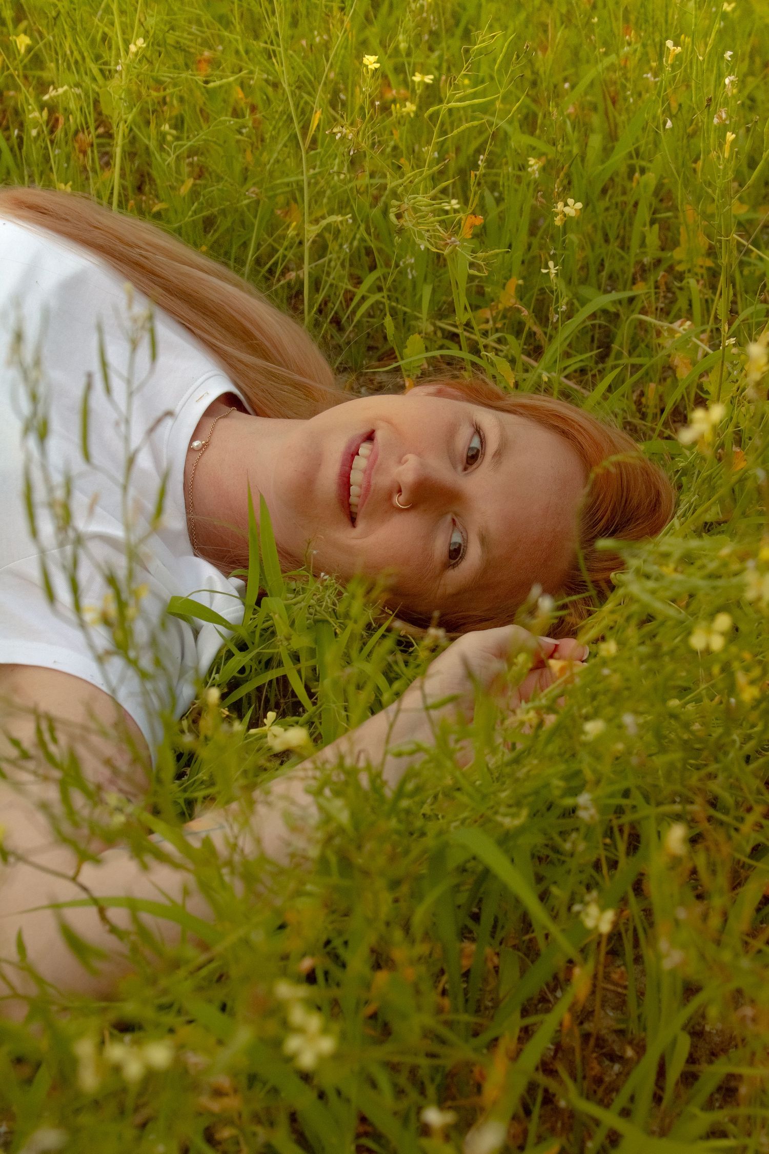 Happy redhead woman lying in tall green grass with wildflowers, smiling at camera in outdoor meadow.