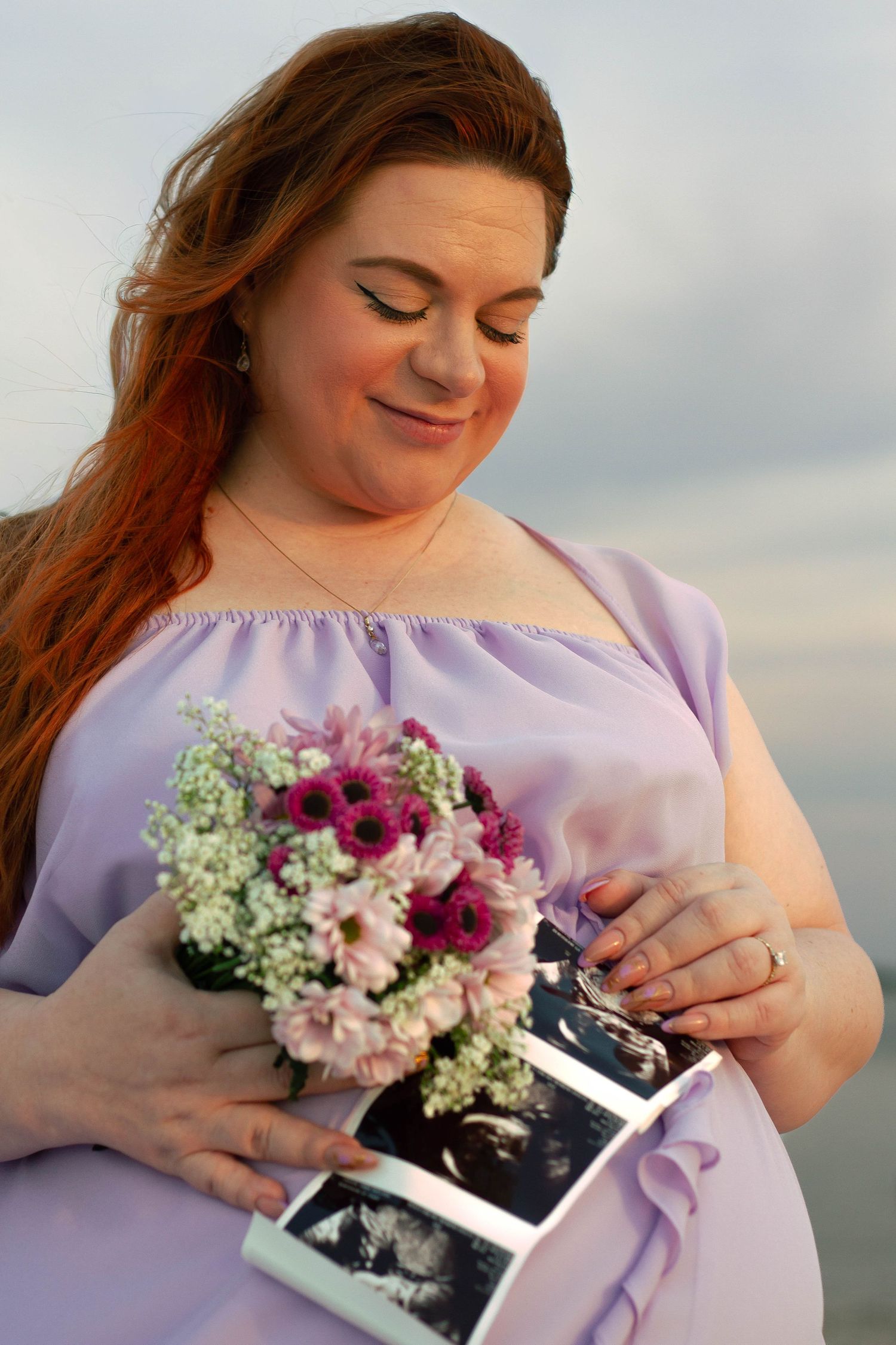 Pregnant woman in lavender dress smiles while holding ultrasound photos and purple flower bouquet.