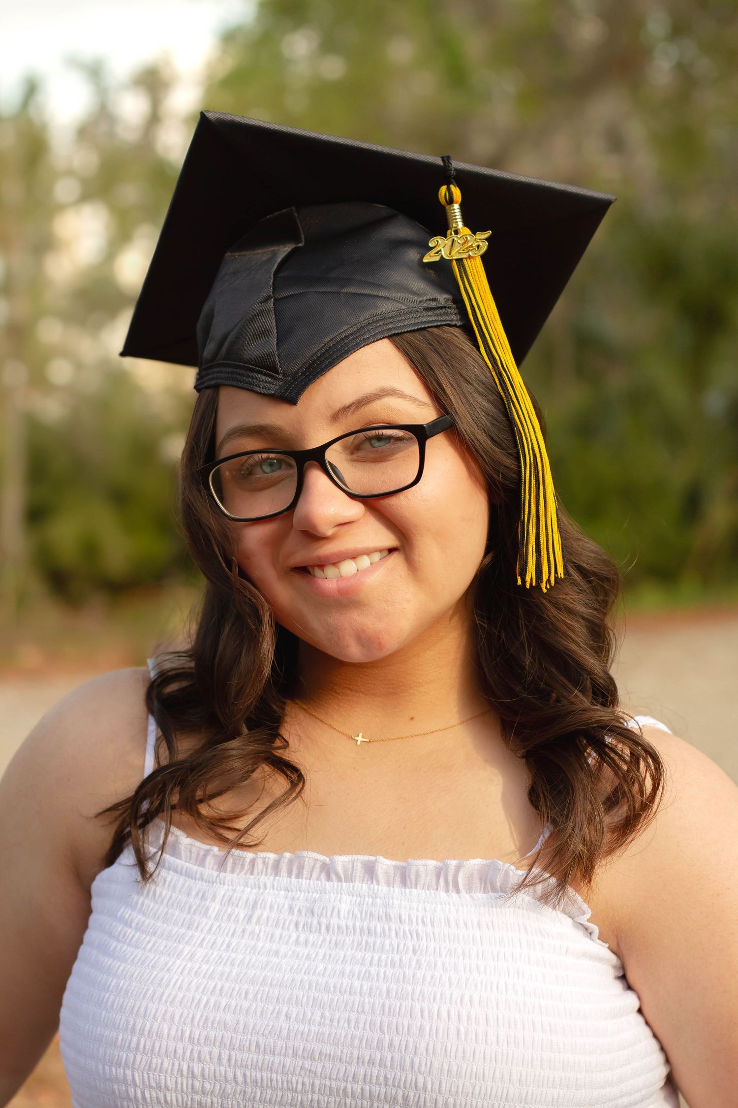 Young woman in black graduation cap with gold tassel and glasses smiling outdoors.
