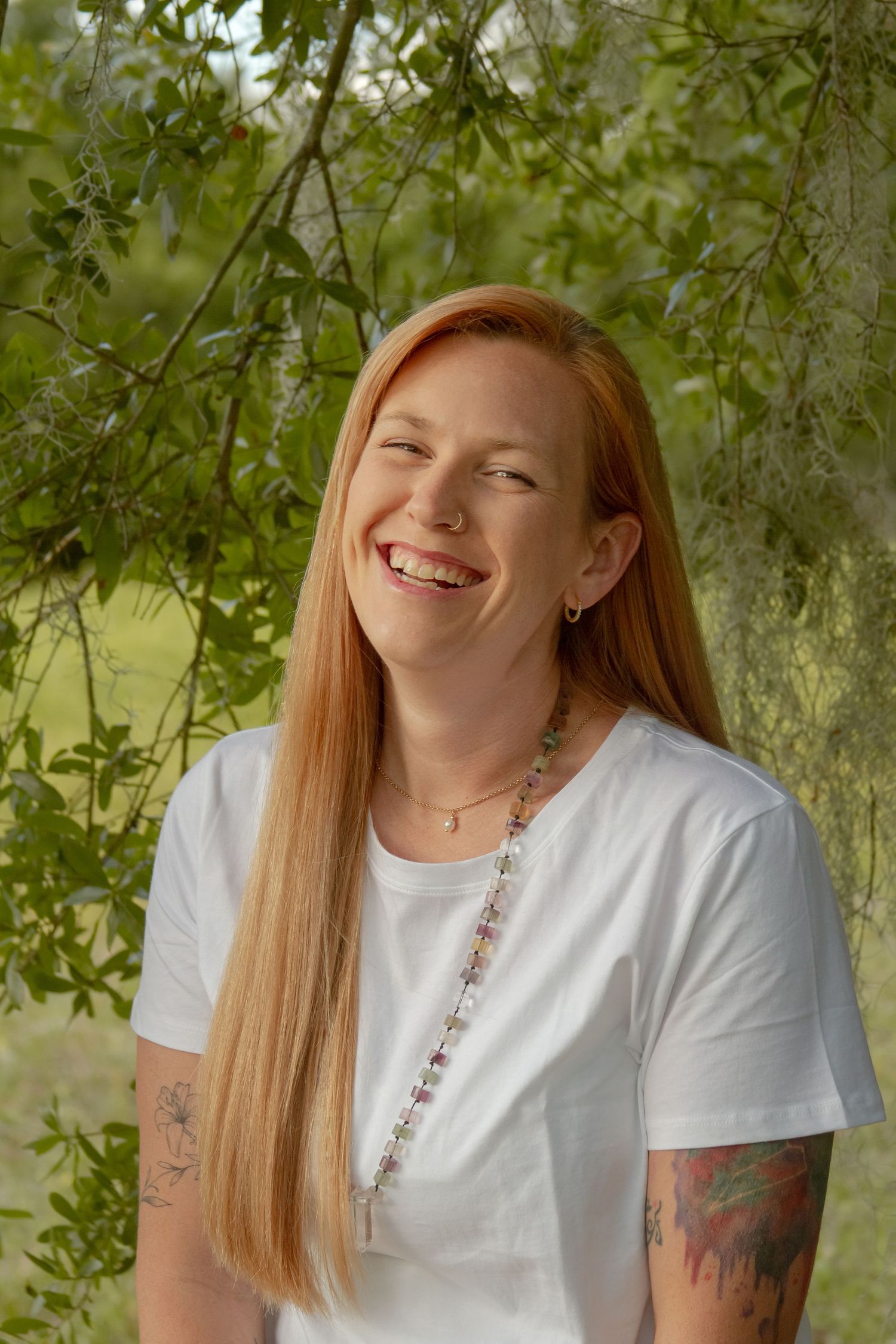 Laughing redhead woman with tattoos in white tee standing under green tree branches outdoors.