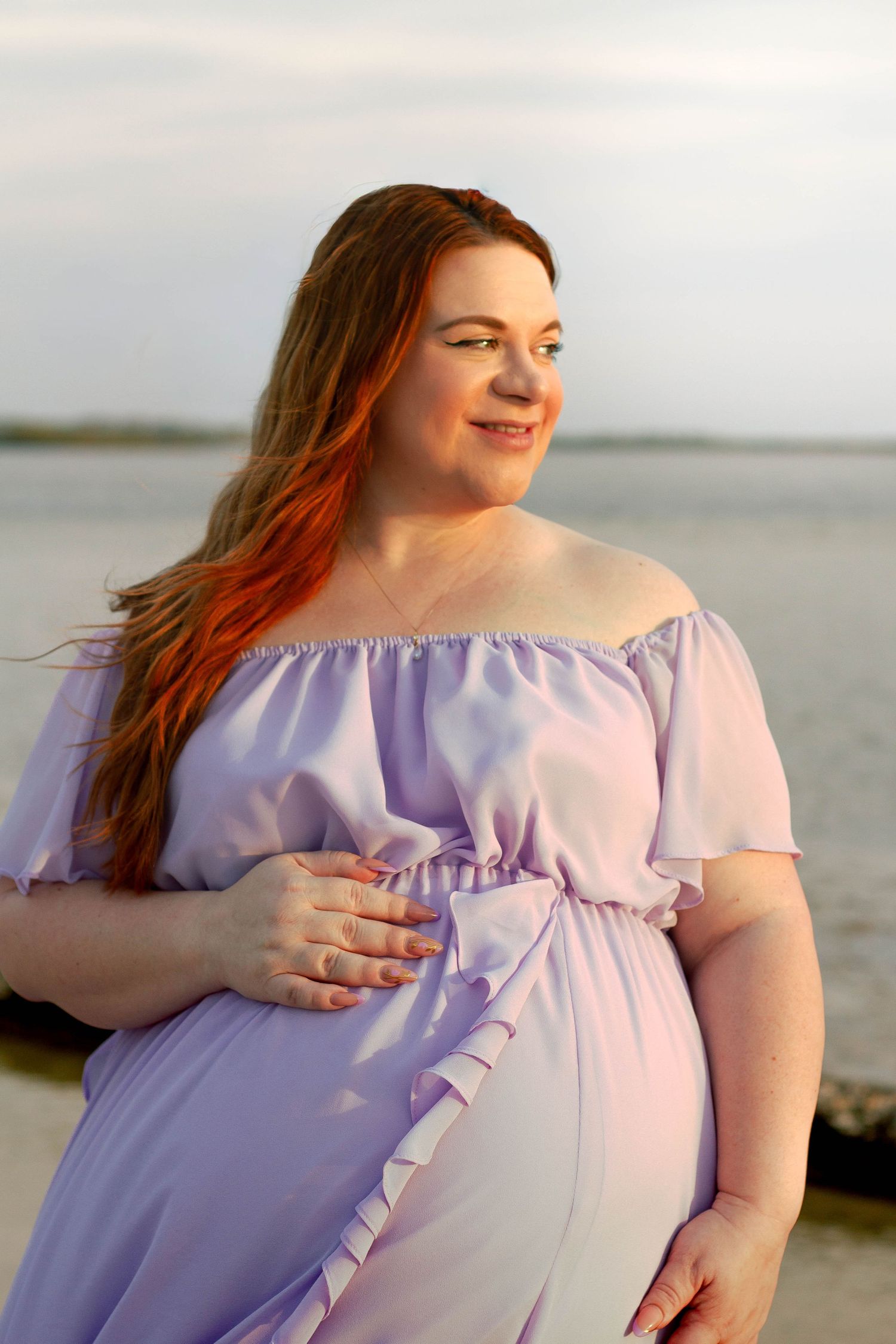 Pregnant redheaded woman in lavender dress gently holds her baby bump while gazing away at the beach.