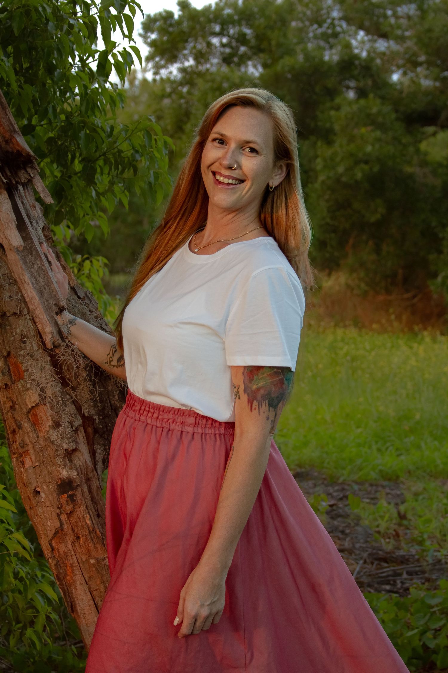 Smiling redhead woman with tattoos in white tee and pink skirt leaning against tree outdoors.