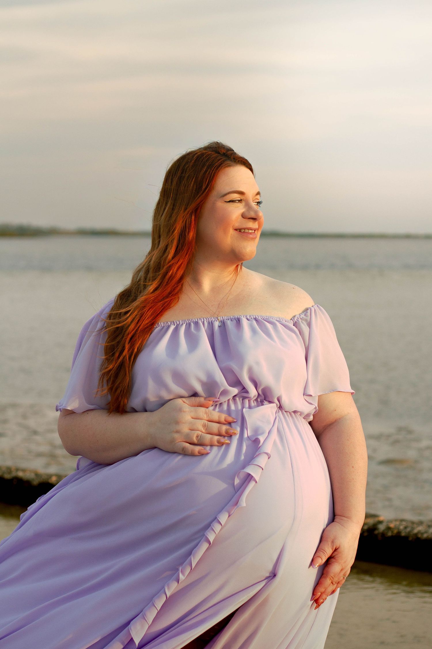 Smiling pregnant woman in off-shoulder lavender dress cradles baby bump by ocean at sunset.