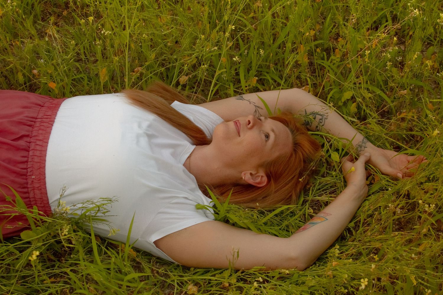 Young redhead woman lying in lush green grass field wearing white t-shirt and pink skirt, relaxing outdoors.