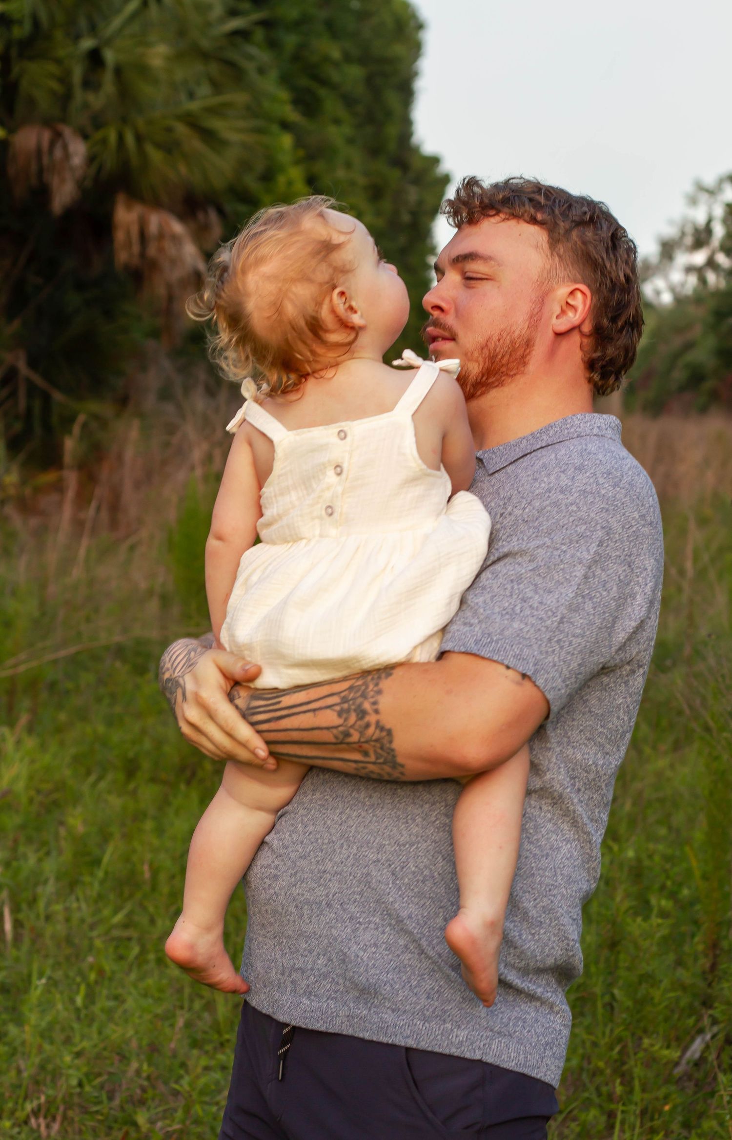 A tattooed father holds his baby girl in a white dress outdoors in a green field, sharing a tender moment.