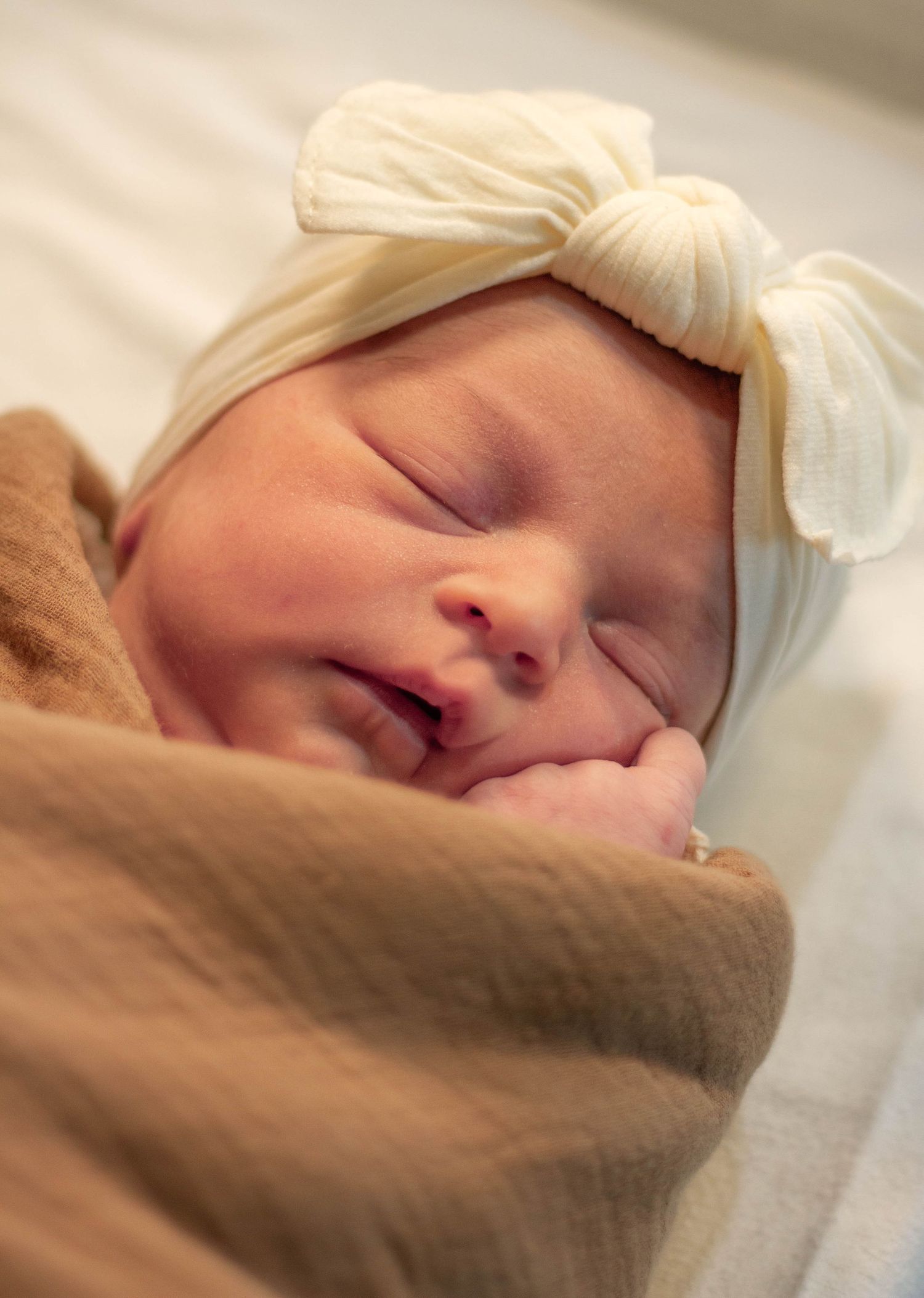 Sleeping newborn baby wrapped in a brown swaddle blanket wearing a white bow hat.