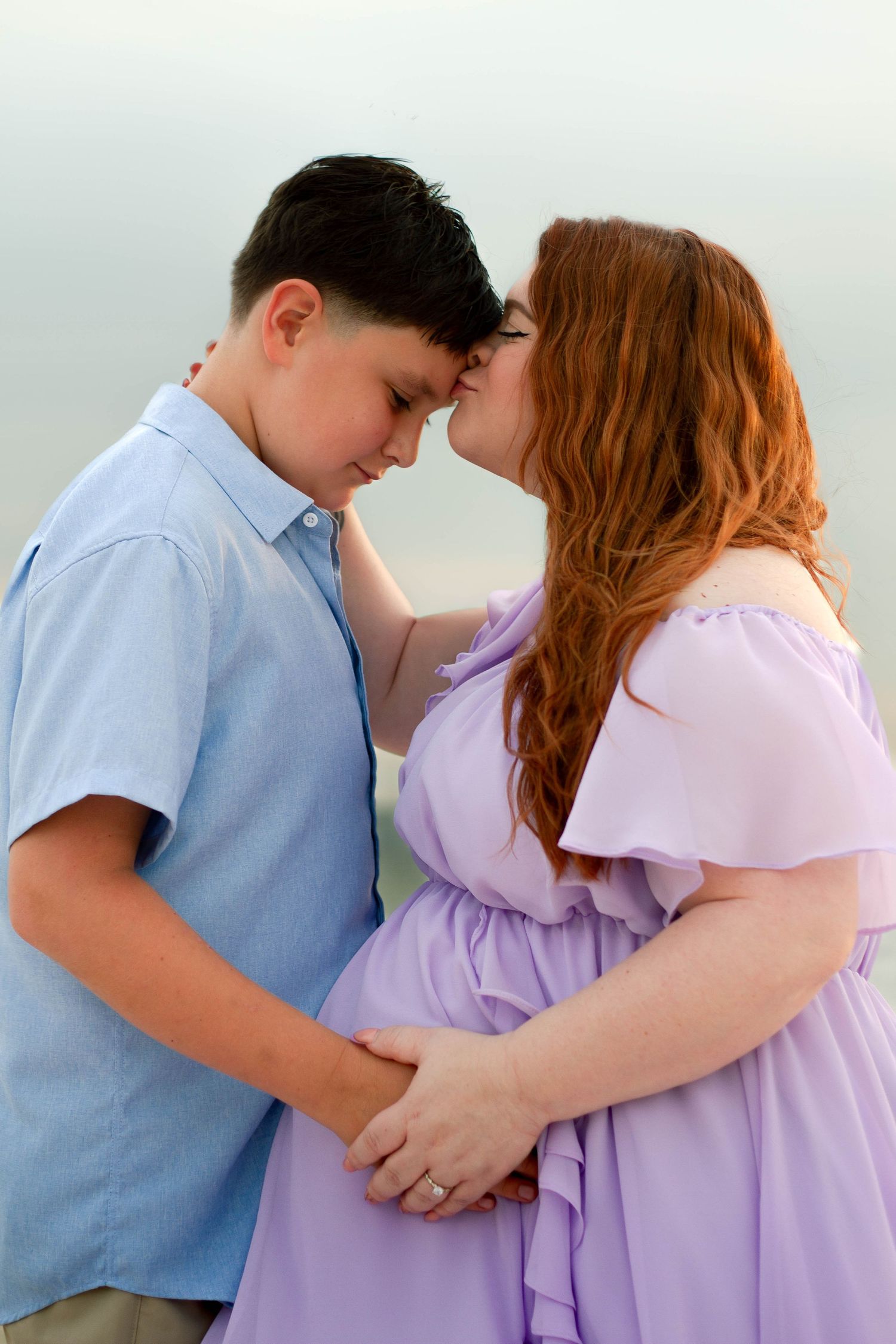 Pregnant woman in lavender dress kisses young sons forehead tenderly while holding hands by the sea.