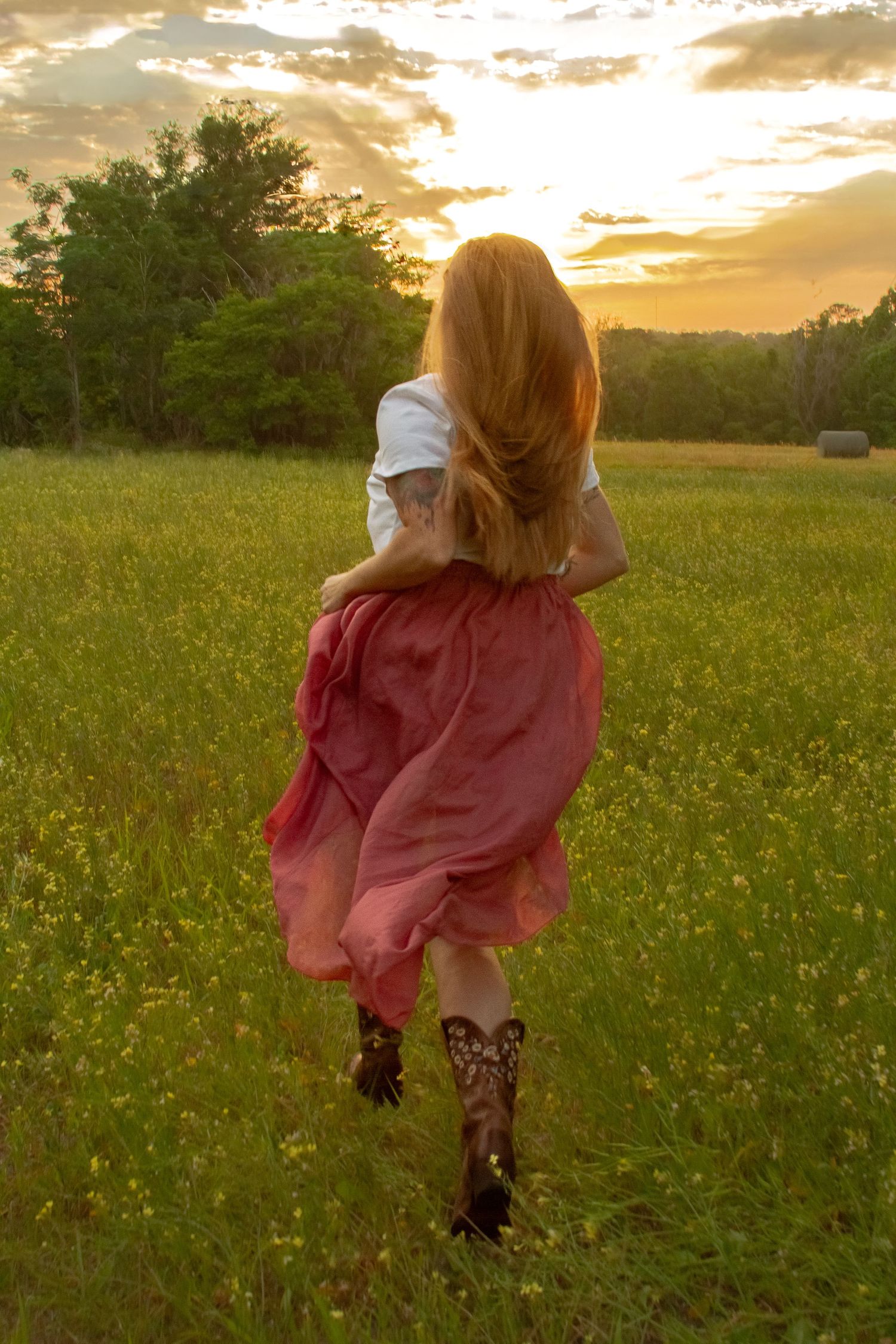 Woman in pink skirt and cowboy boots running through golden sunset meadow.