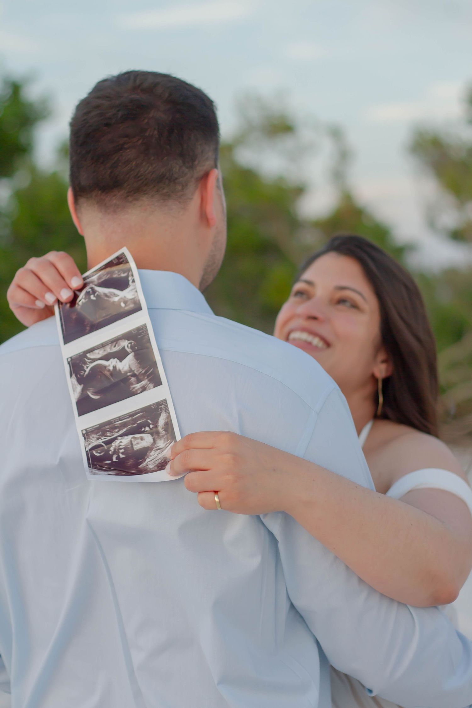 Happy pregnant couple on beach, woman smiling while holding ultrasound photos against partner's back.