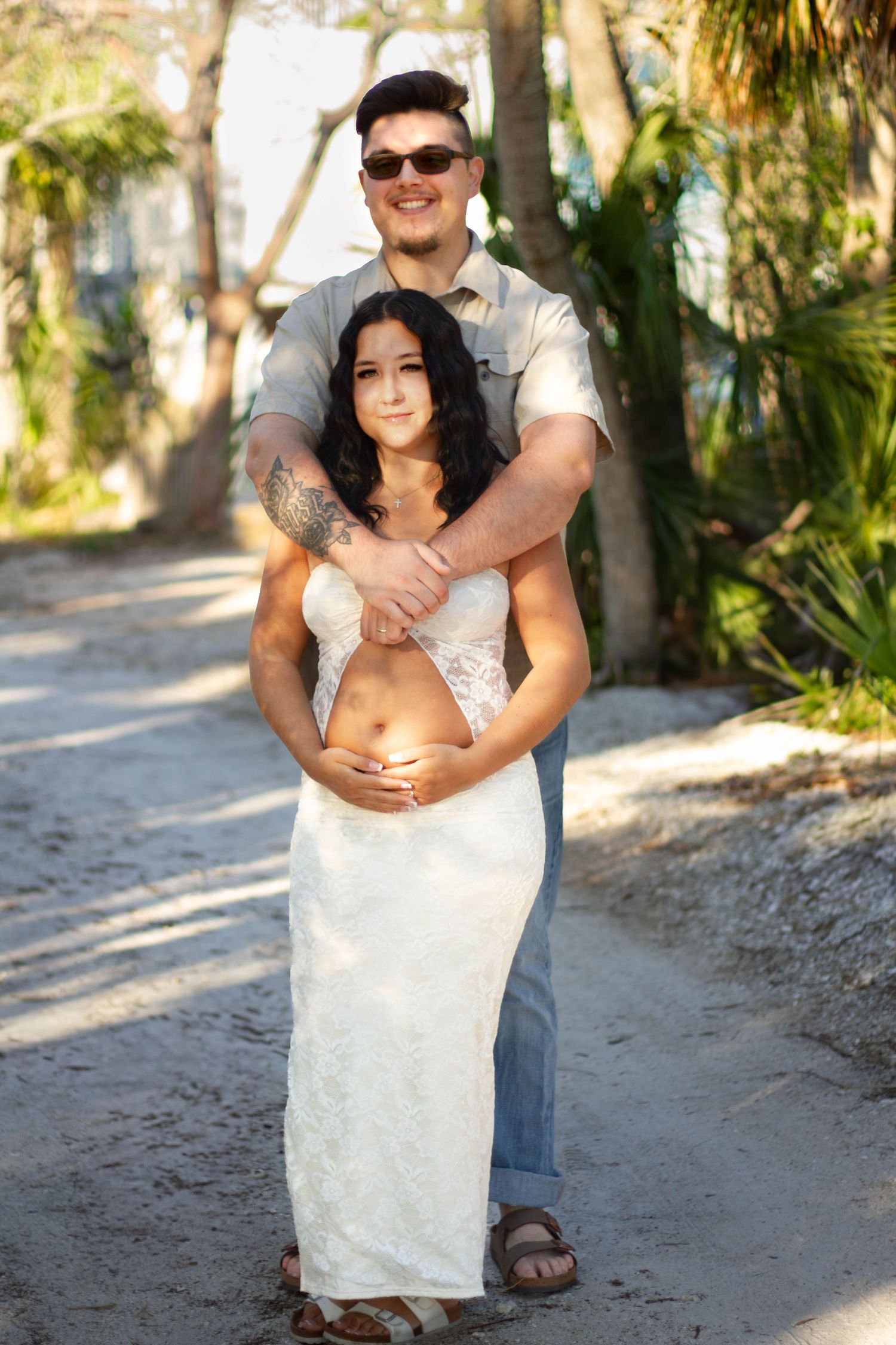 Man embracing pregnant woman from behind on sandy path surrounded by palm trees.