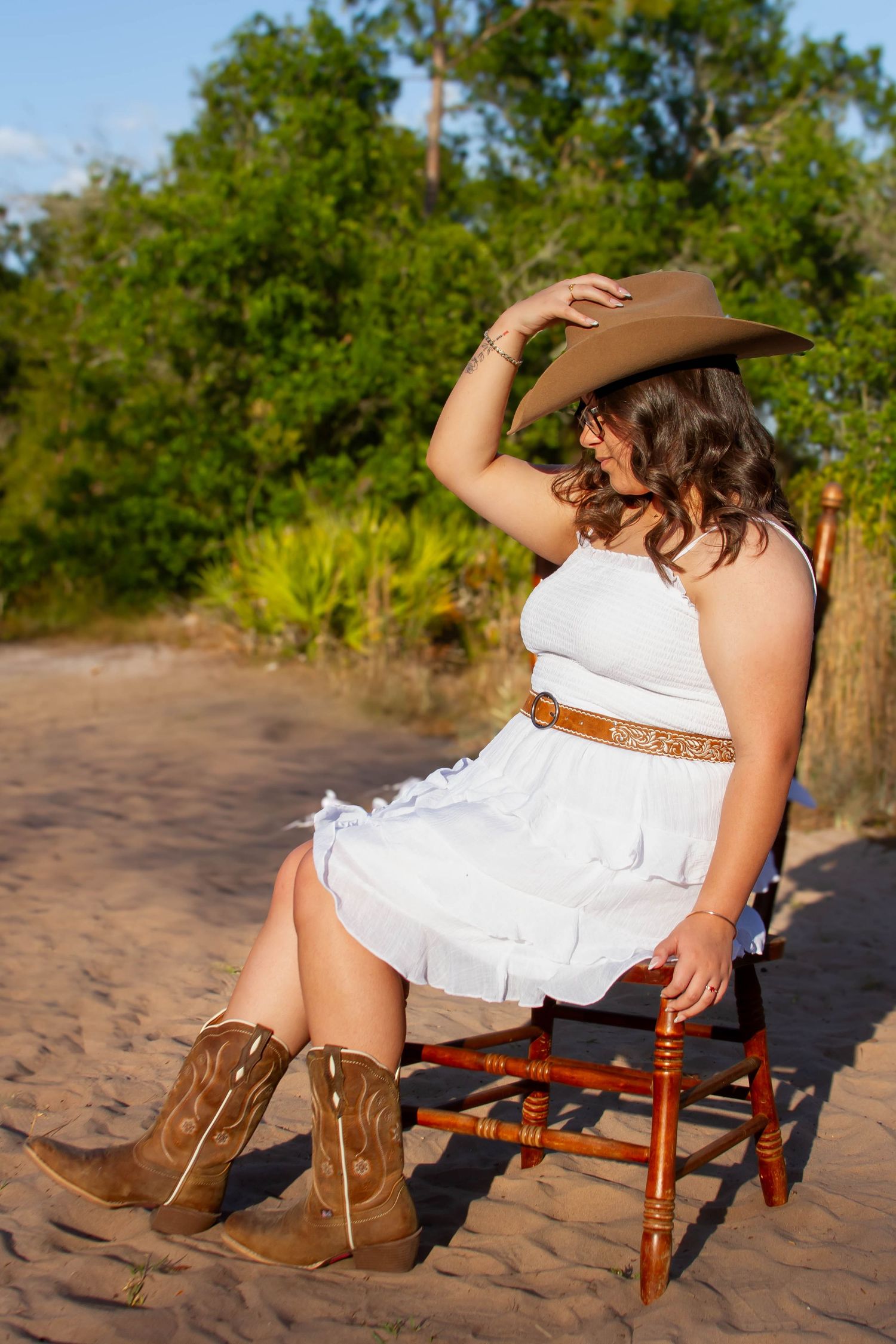 Woman in white dress, cowboy hat and boots sitting on wooden chair in sandy outdoor setting.