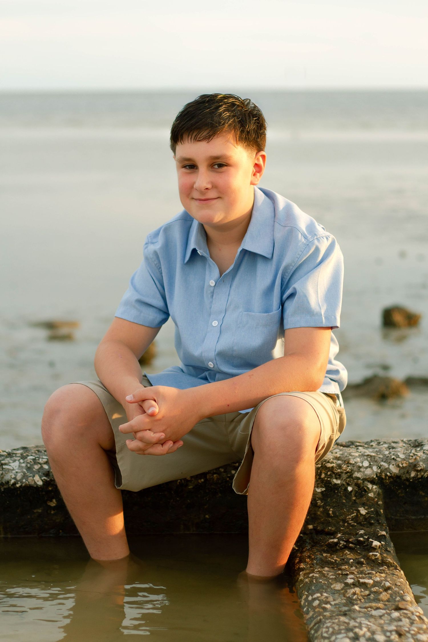 Smiling preteen boy in blue shirt sits on a rock jetty with feet in the water at sunset.