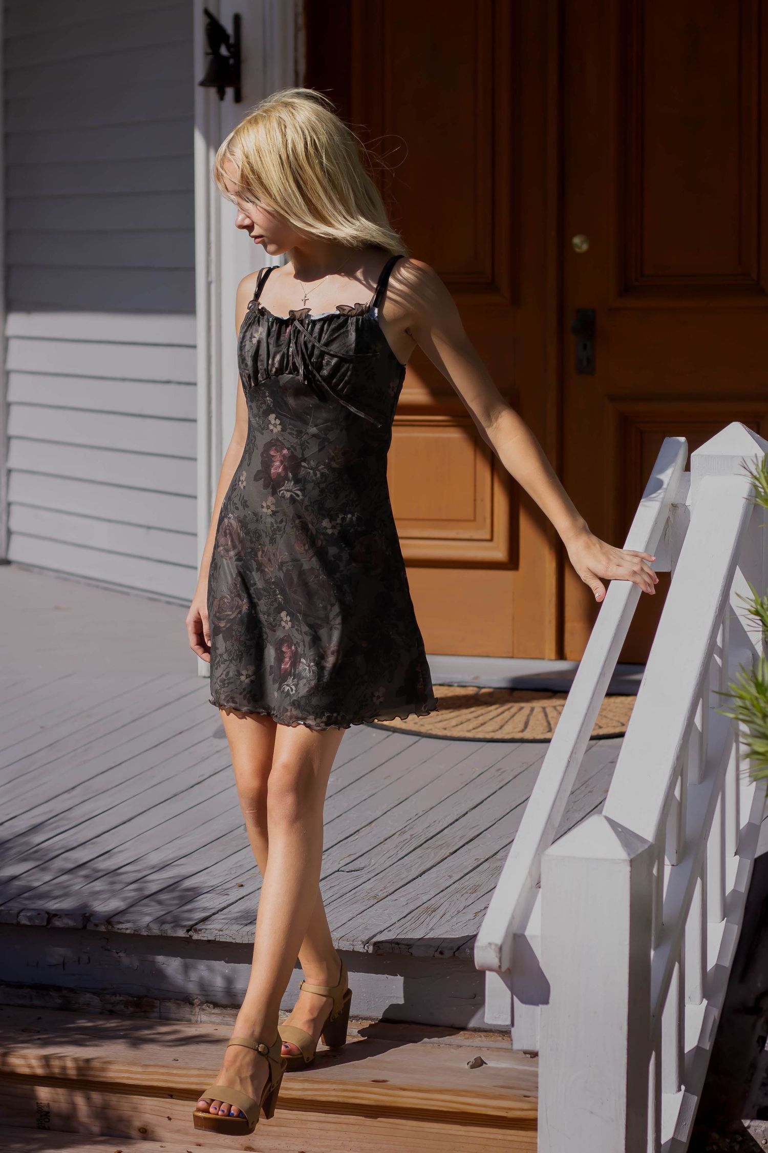 Blonde woman in dark floral dress and wooden heels descending sunny porch steps by a brown door.