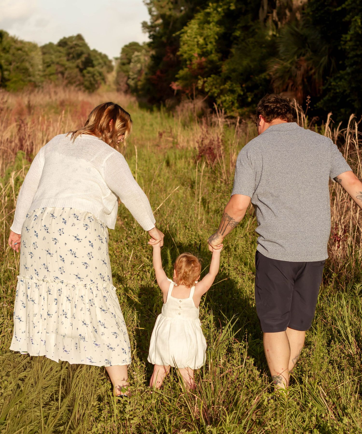 Family of three walking through a lush green meadow, parents holding toddler's hands from behind.