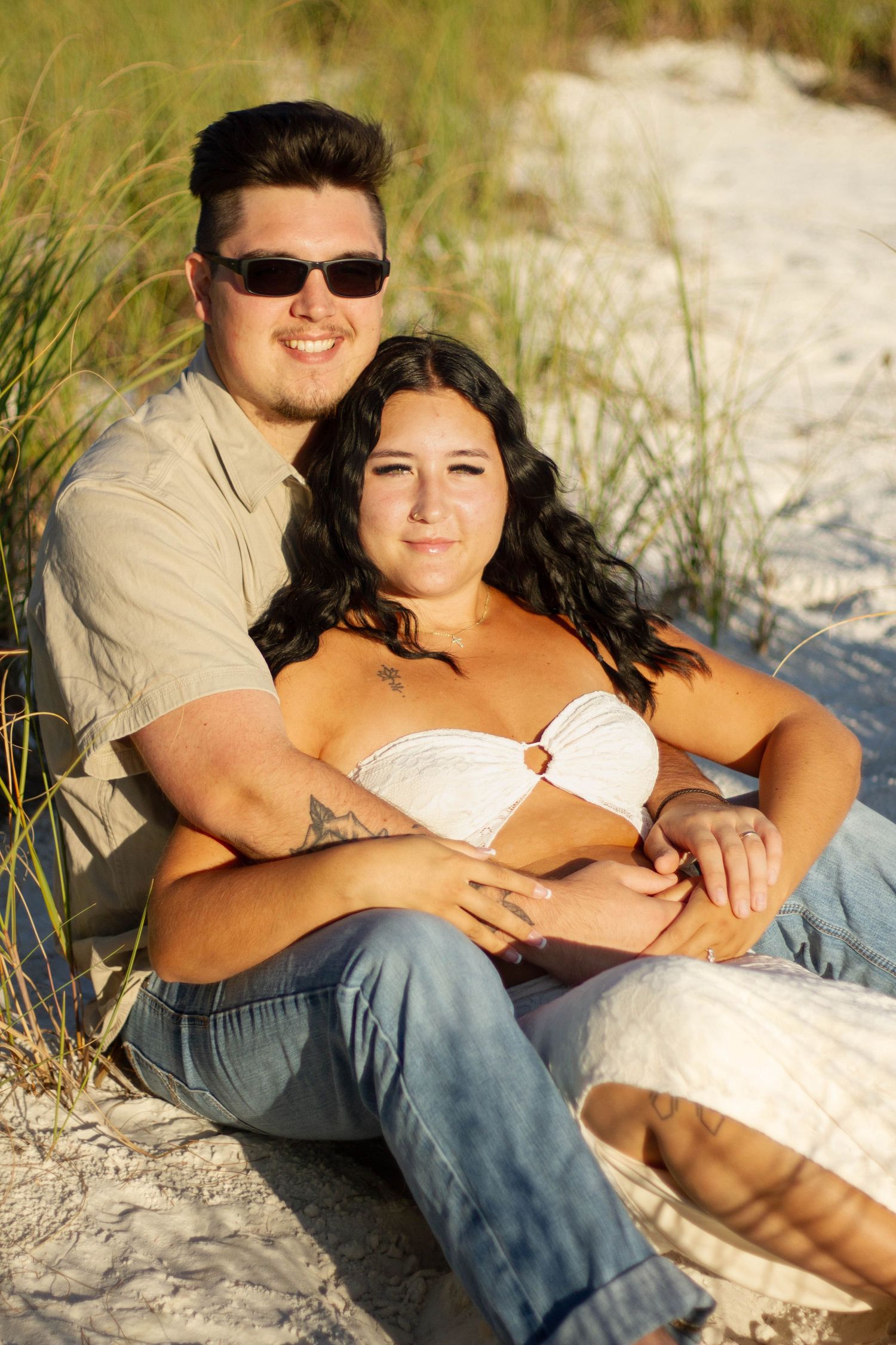 Couple sitting together in beach grass, man in sunglasses embracing pregnant partner.