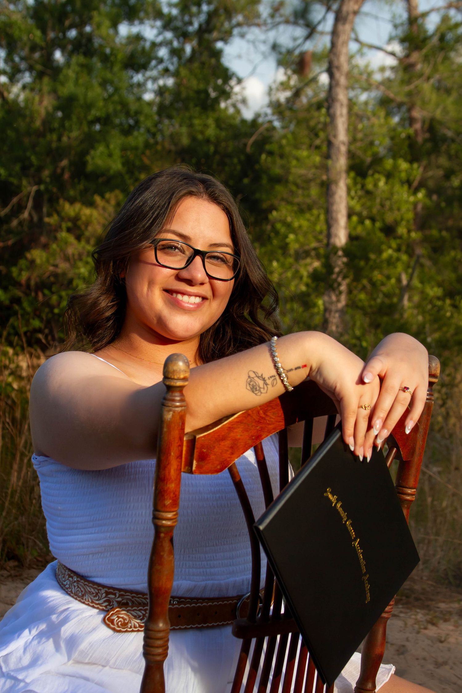 Smiling young woman with glasses holding diploma, seated on wooden chair in outdoor nature setting.
