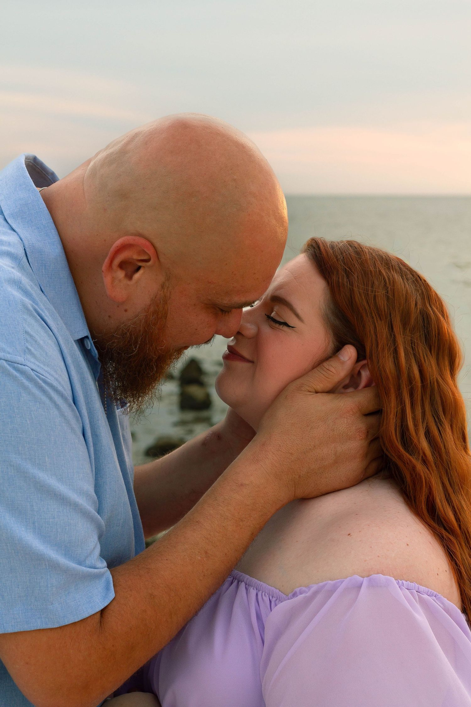 Bearded man tenderly holds pregnant redheaded woman's face as they touch noses on the beach.