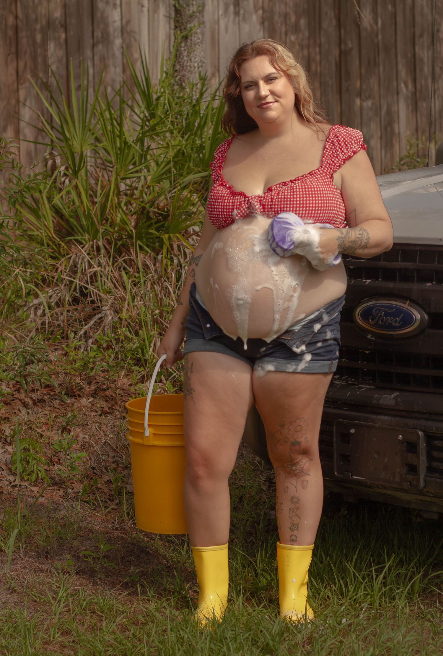 A pregnant woman in a red gingham top and yellow rubber boots washes a Ford truck with a soapy sponge.
