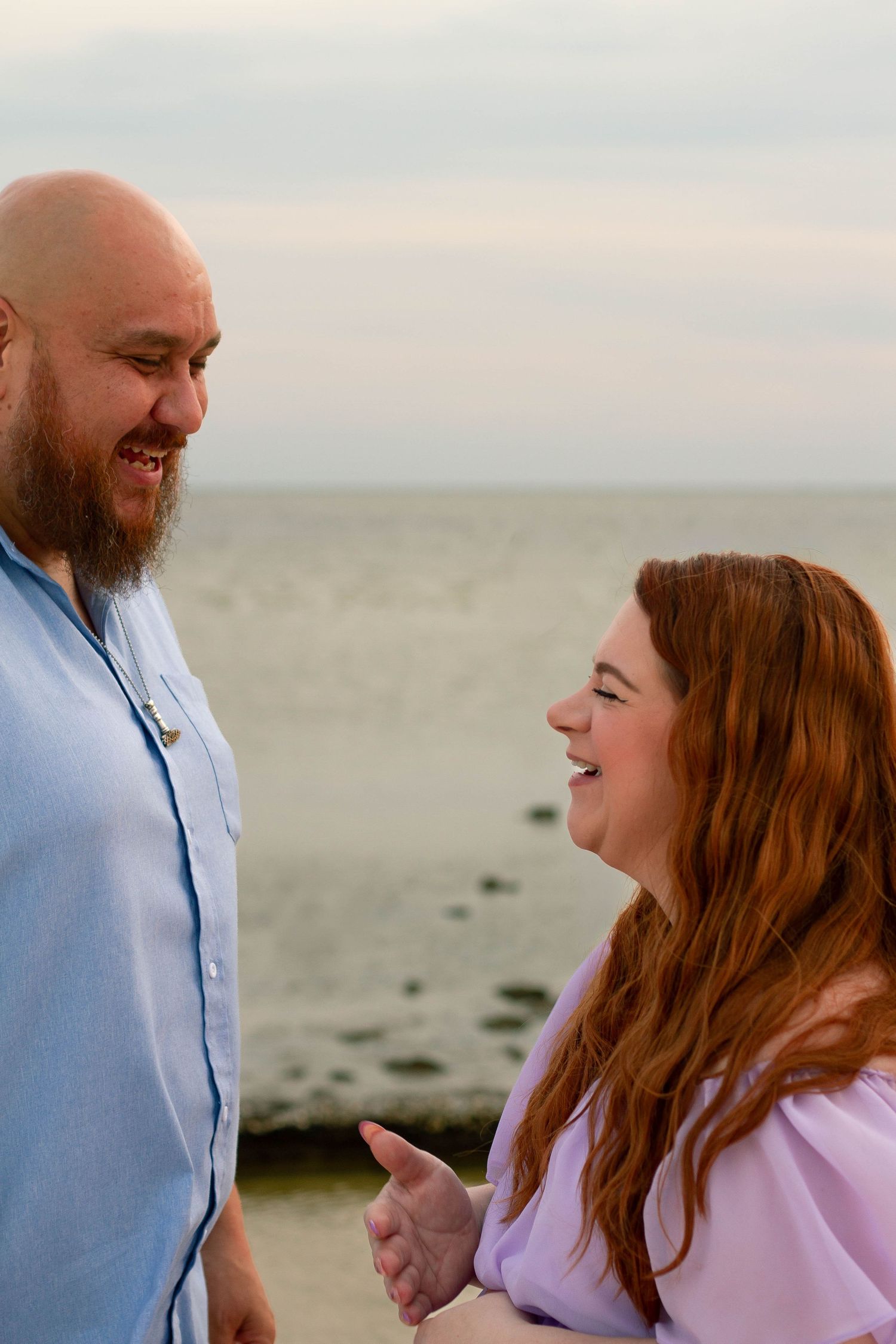 Bearded man and redheaded pregnant woman laugh together during a beach maternity photo session.