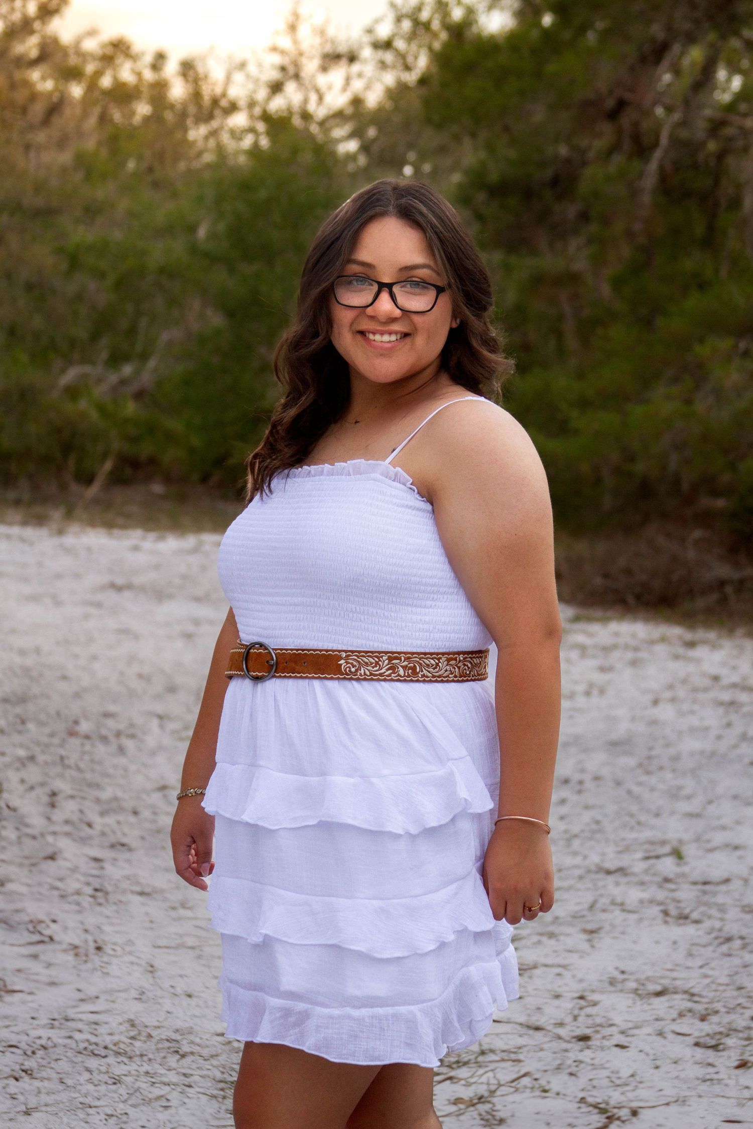 Smiling young woman in white ruffled dress with brown belt standing on sandy path.