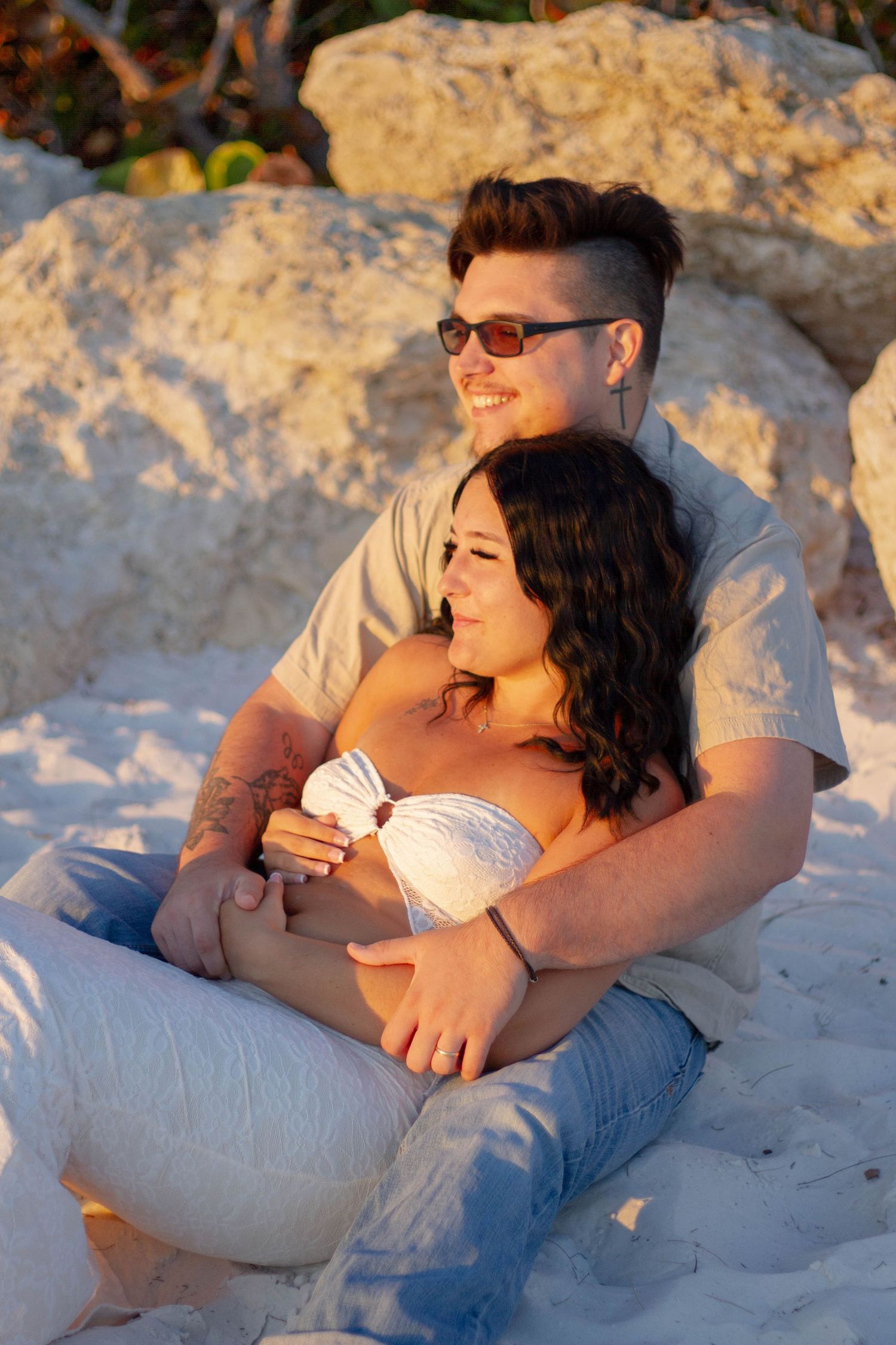 Happy expecting couple sitting against sandy rocks at sunset, man embracing pregnant woman warmly.
