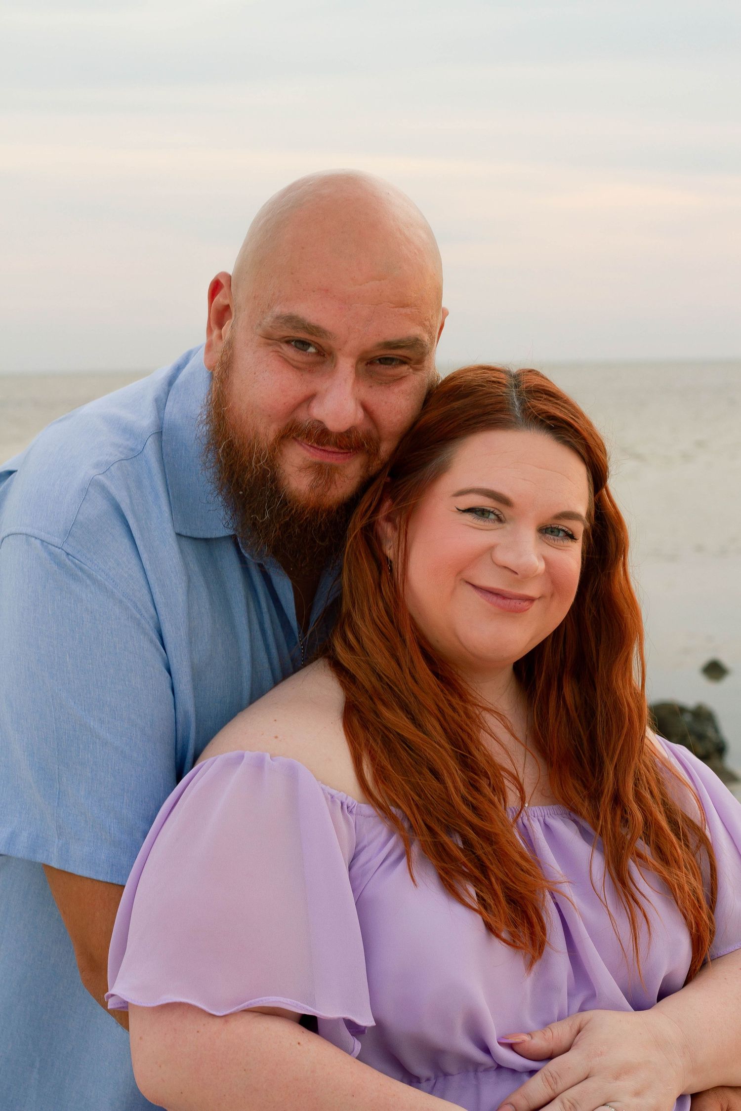 Bearded man embraces smiling redhead pregnant woman during beach maternity photo session.