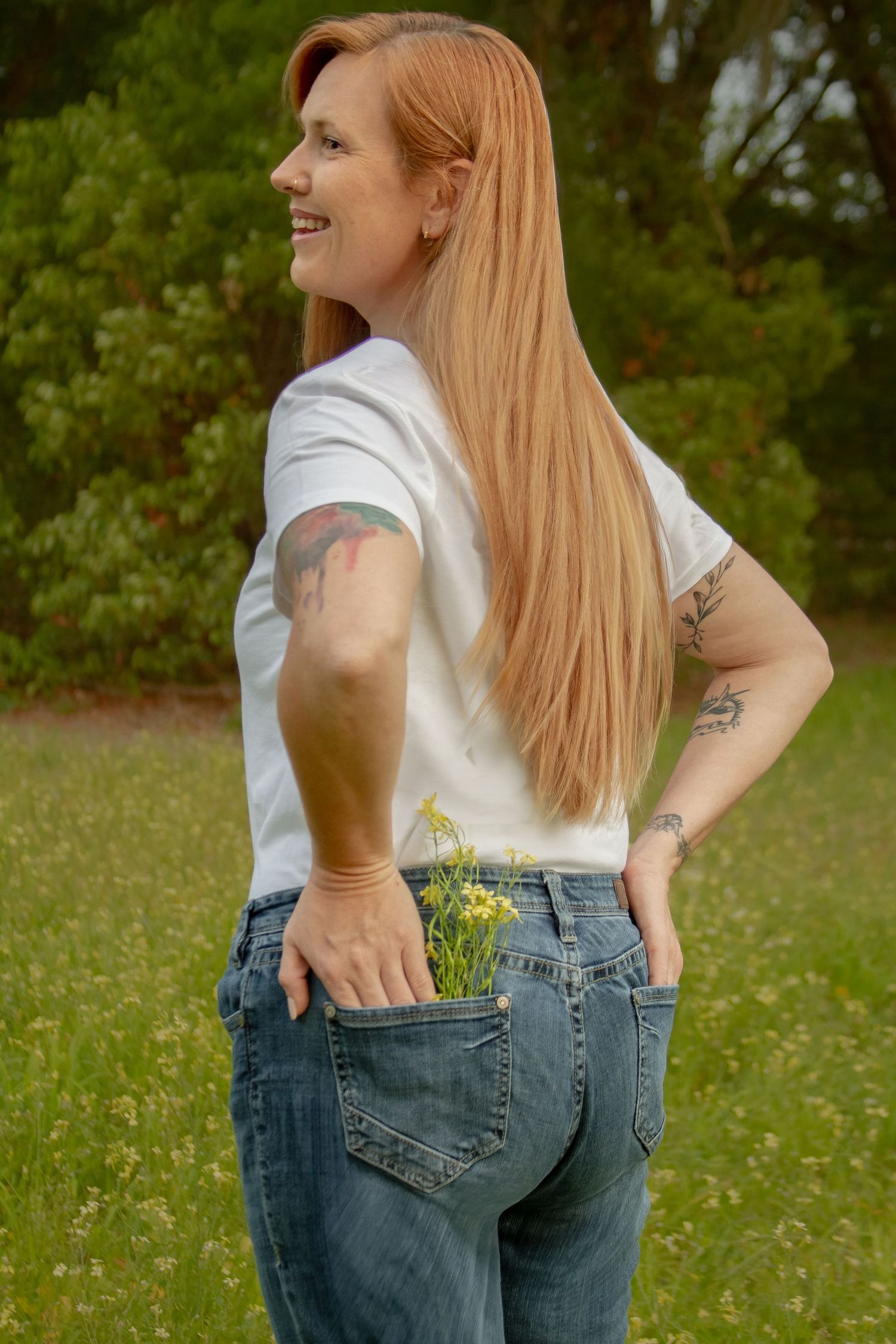 Redhead woman in white t-shirt and jeans with wildflowers in pocket, showing tattoos in green field.