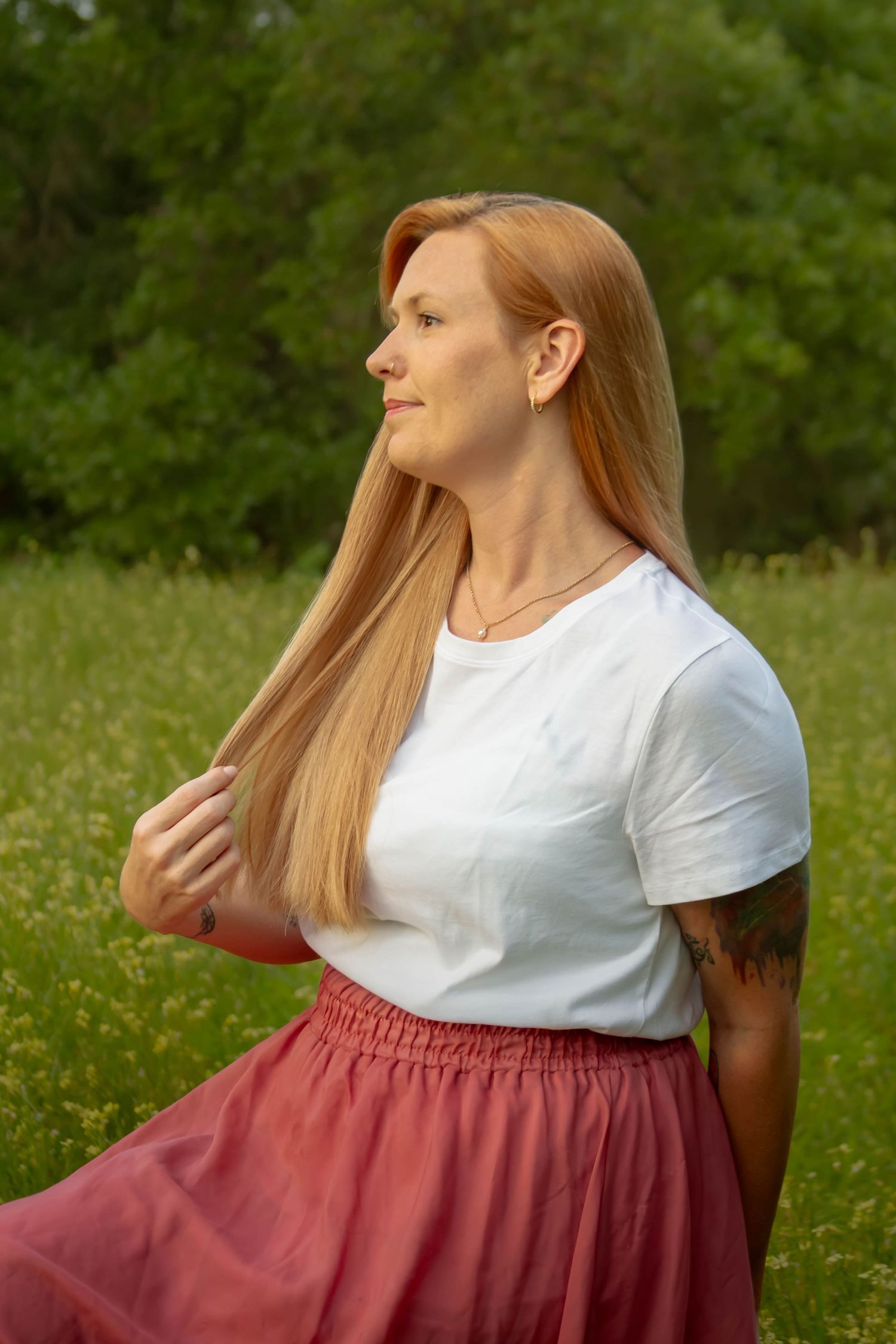 Redhead woman in white t-shirt and pink skirt holding her hair while gazing sideways in a meadow.