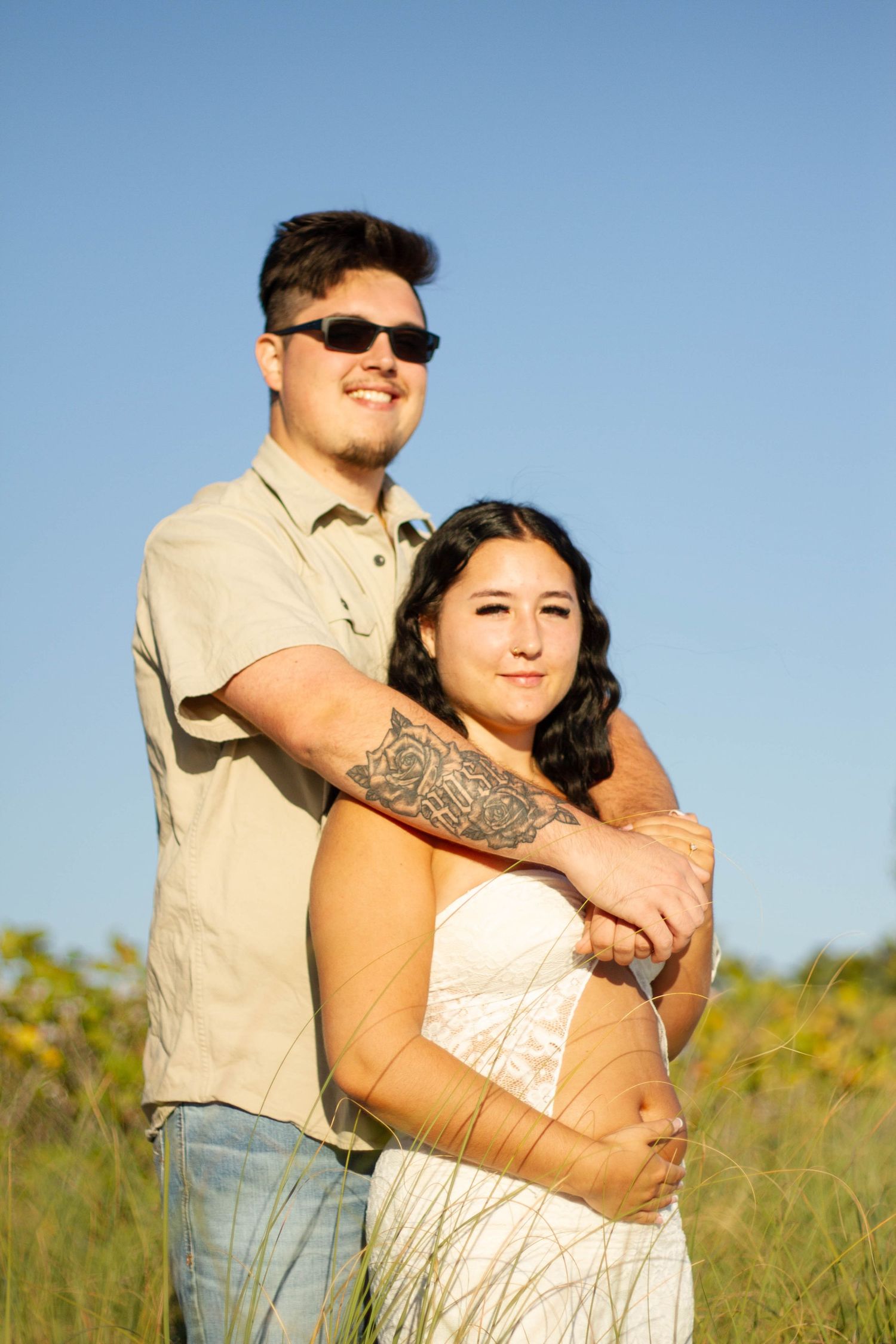 Man in sunglasses embracing pregnant woman in white dress in sunny field.