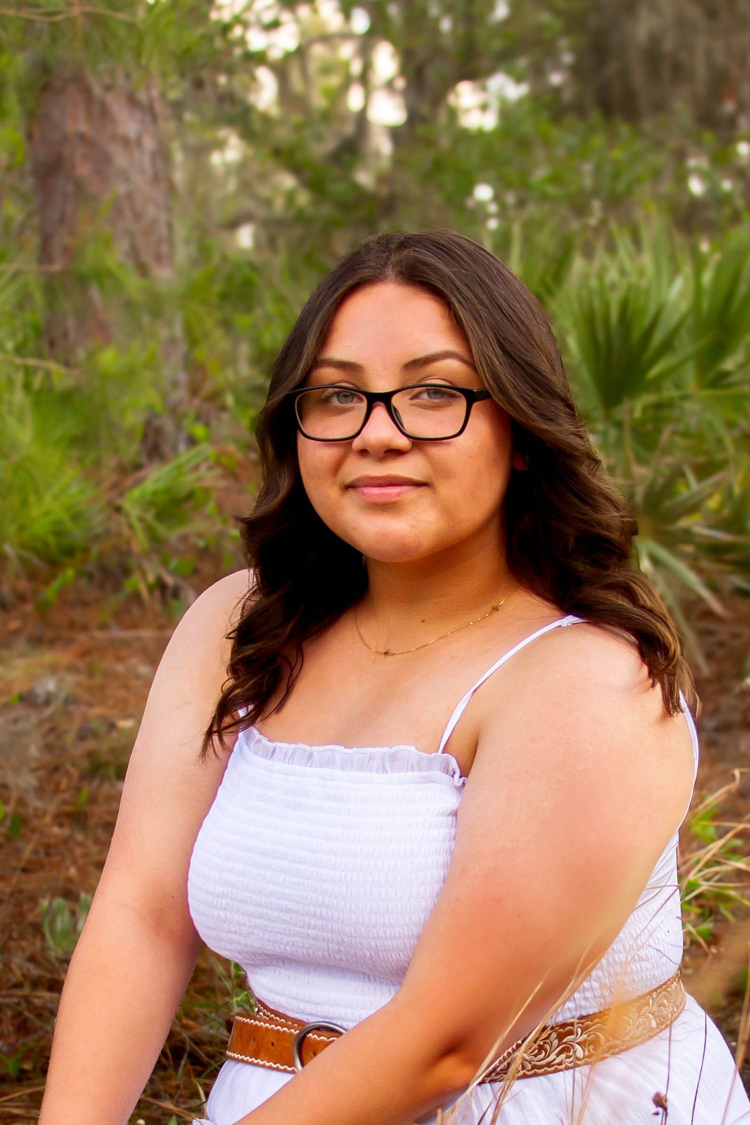 Young woman with glasses and wavy hair wearing white dress with brown belt in forest setting.