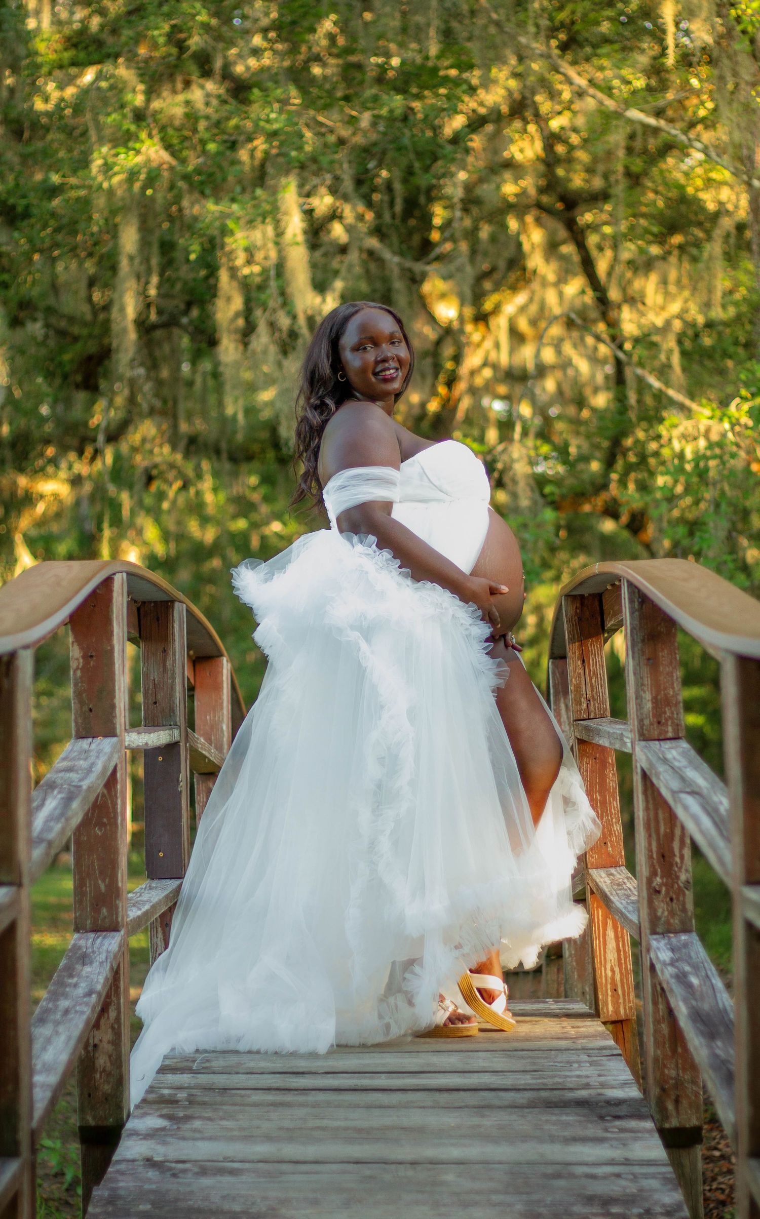 Pregnant woman in white off-shoulder tulle gown posing on wooden bridge surrounded by lush green trees.