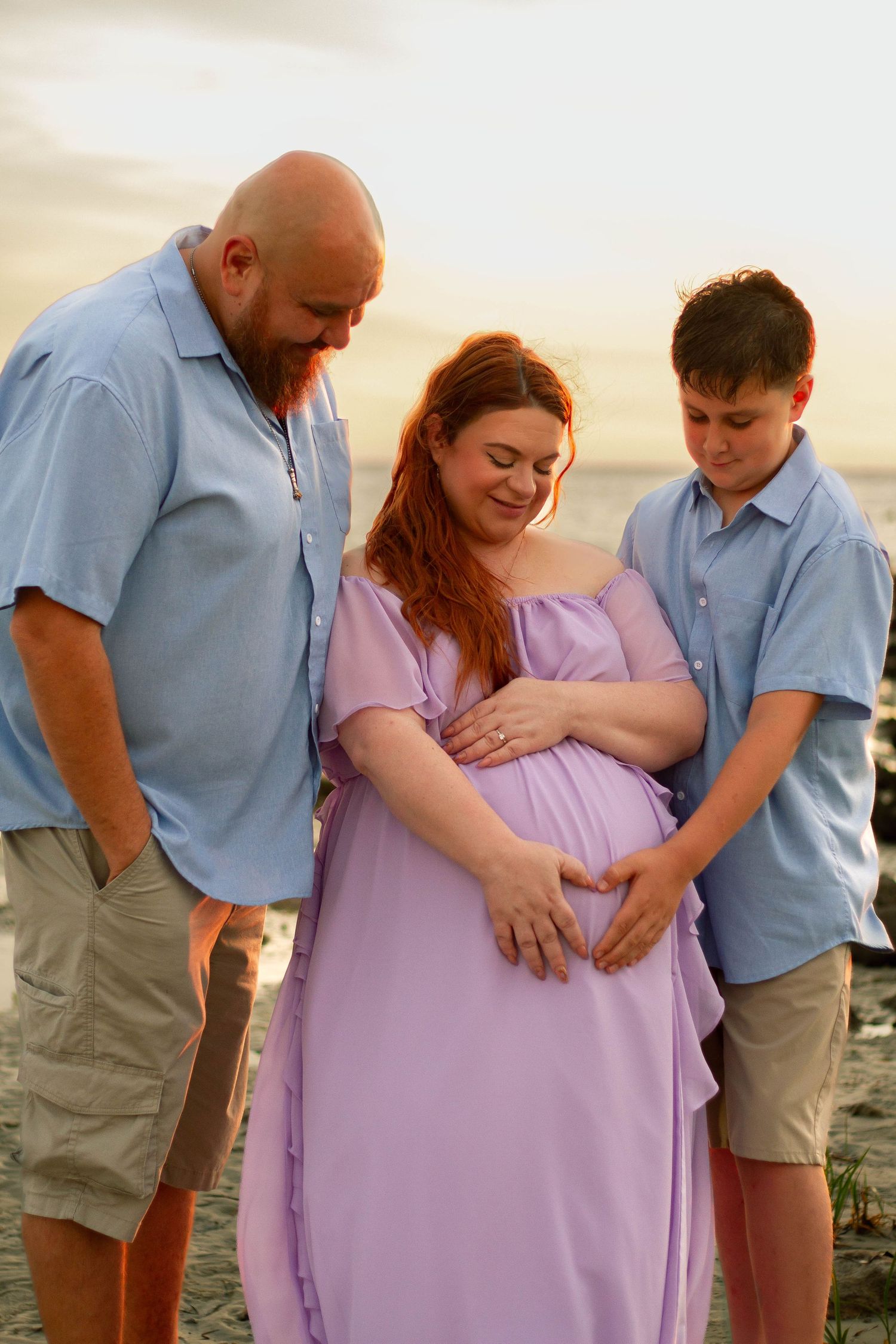 A family maternity beach photo at golden hour with a father, pregnant mother, and teenage son.