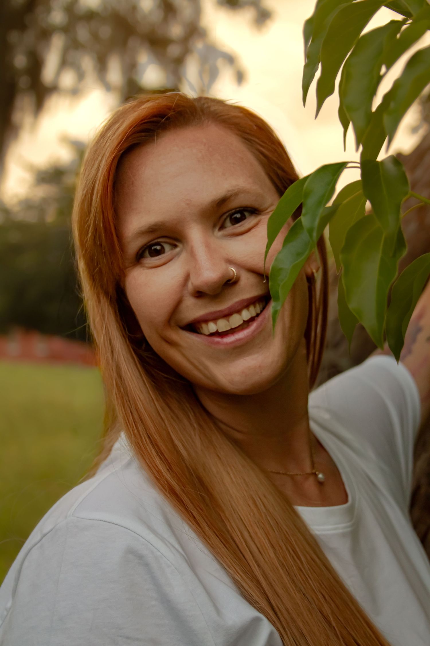 Close-up portrait of a smiling redheaded woman with a nose ring beside green leaves at sunset.