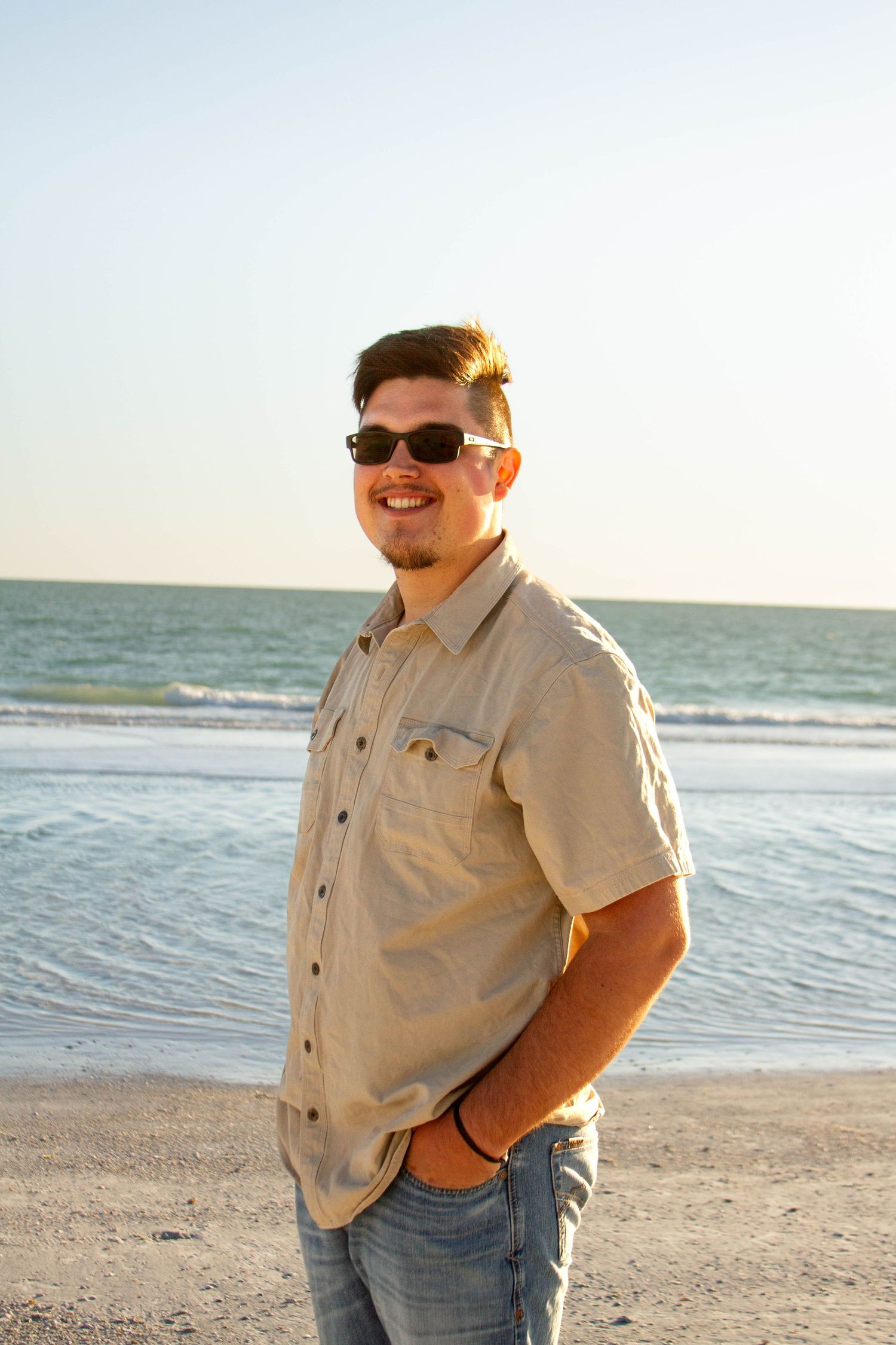 Smiling man in sunglasses and beige shirt standing on sandy beach at sunset.