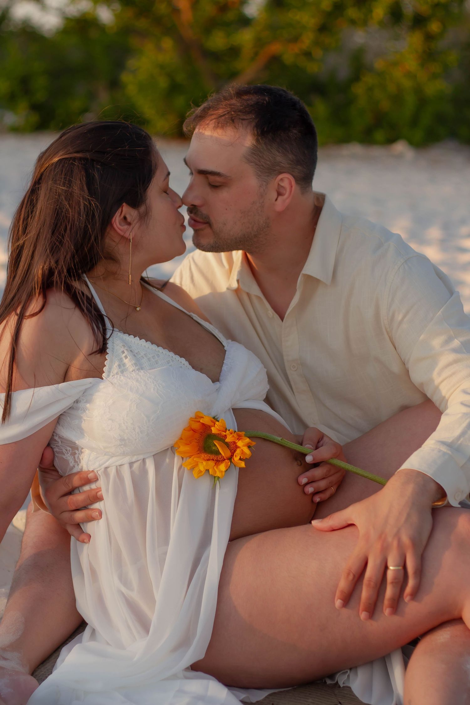 Couple nearly kissing on sandy beach at sunset, woman in white outfit holding a sunflower on her bare belly.