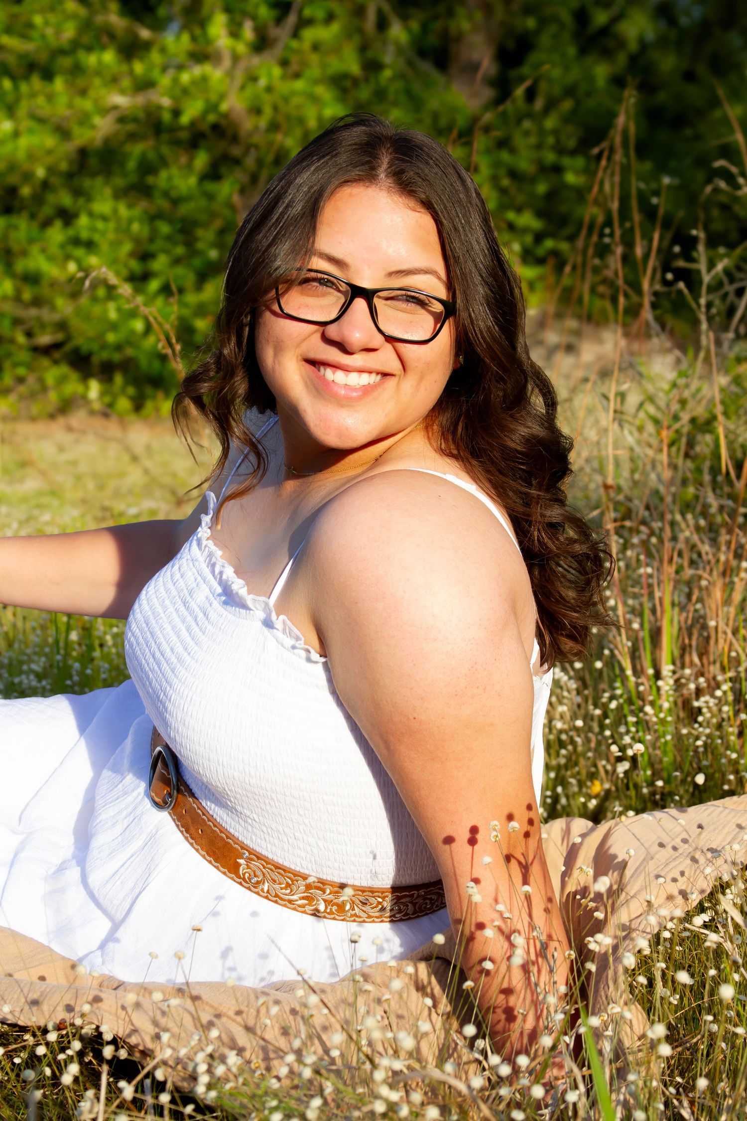 Young woman in white dress with brown belt smiling while sitting in wildflower meadow.