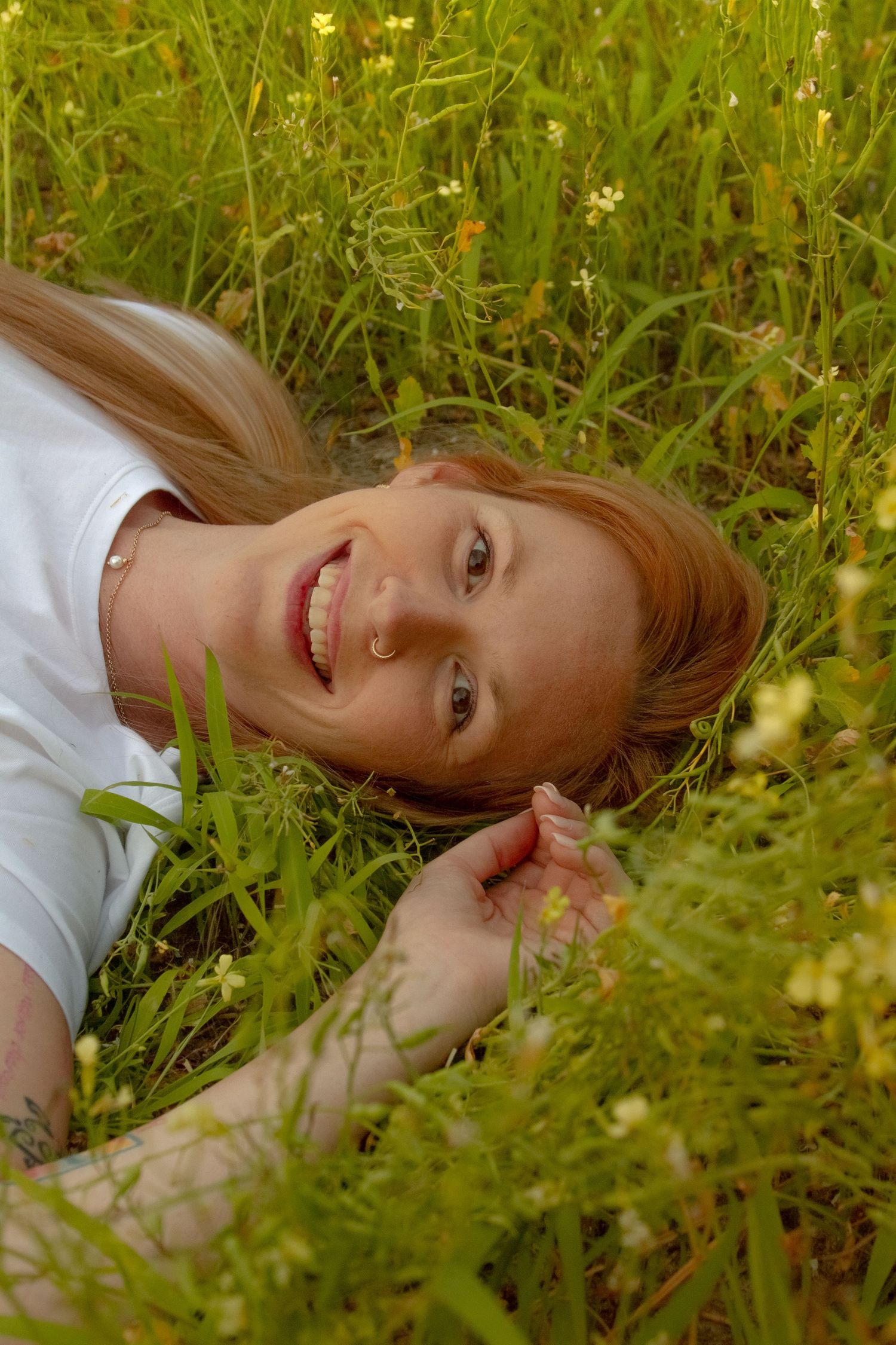 Smiling redhead woman lying in wildflower meadow, surrounded by green grass and small white flowers.