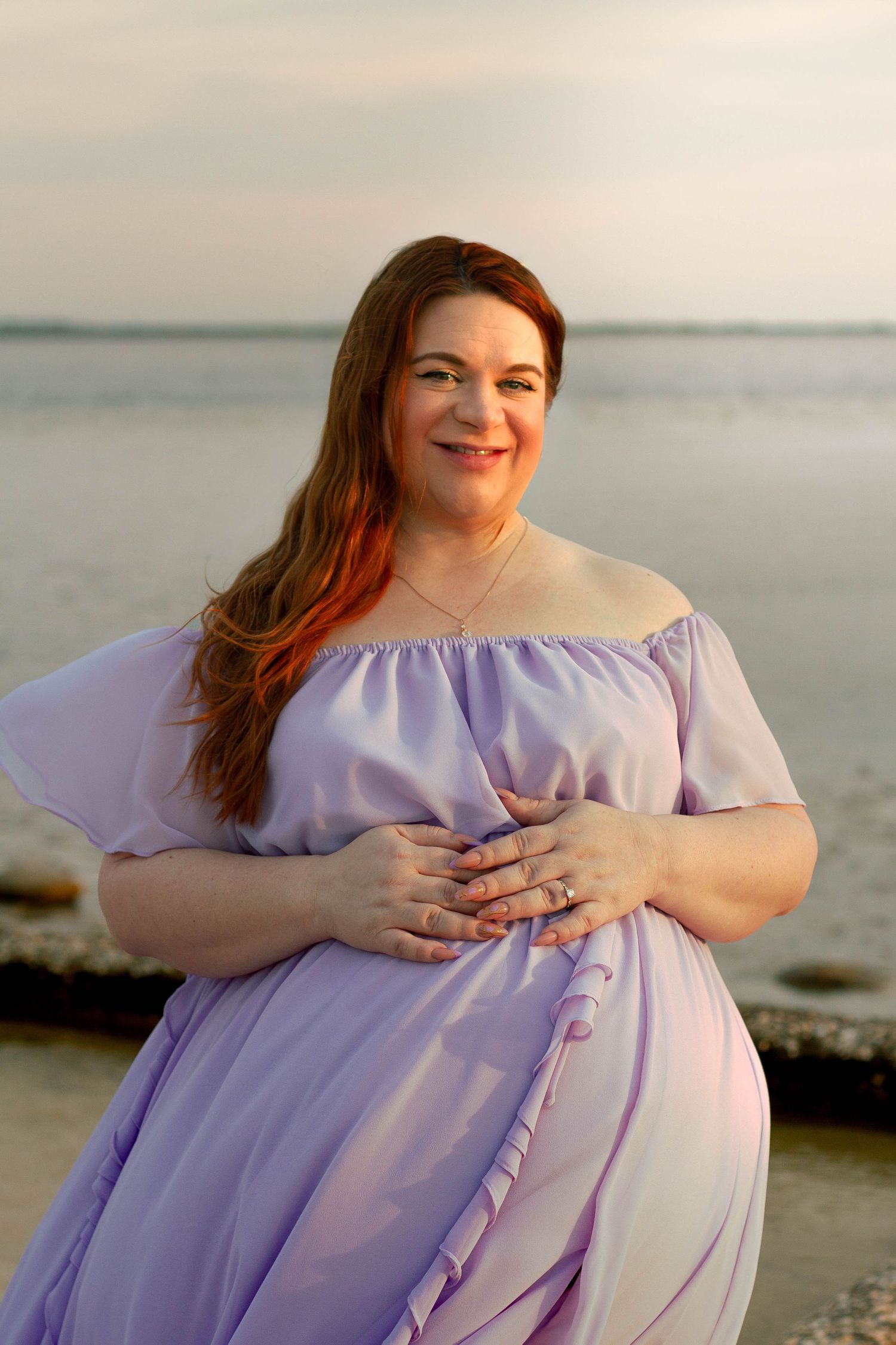 Happy pregnant  woman in lavender off-shoulder dress holds baby bump on rocky beach.