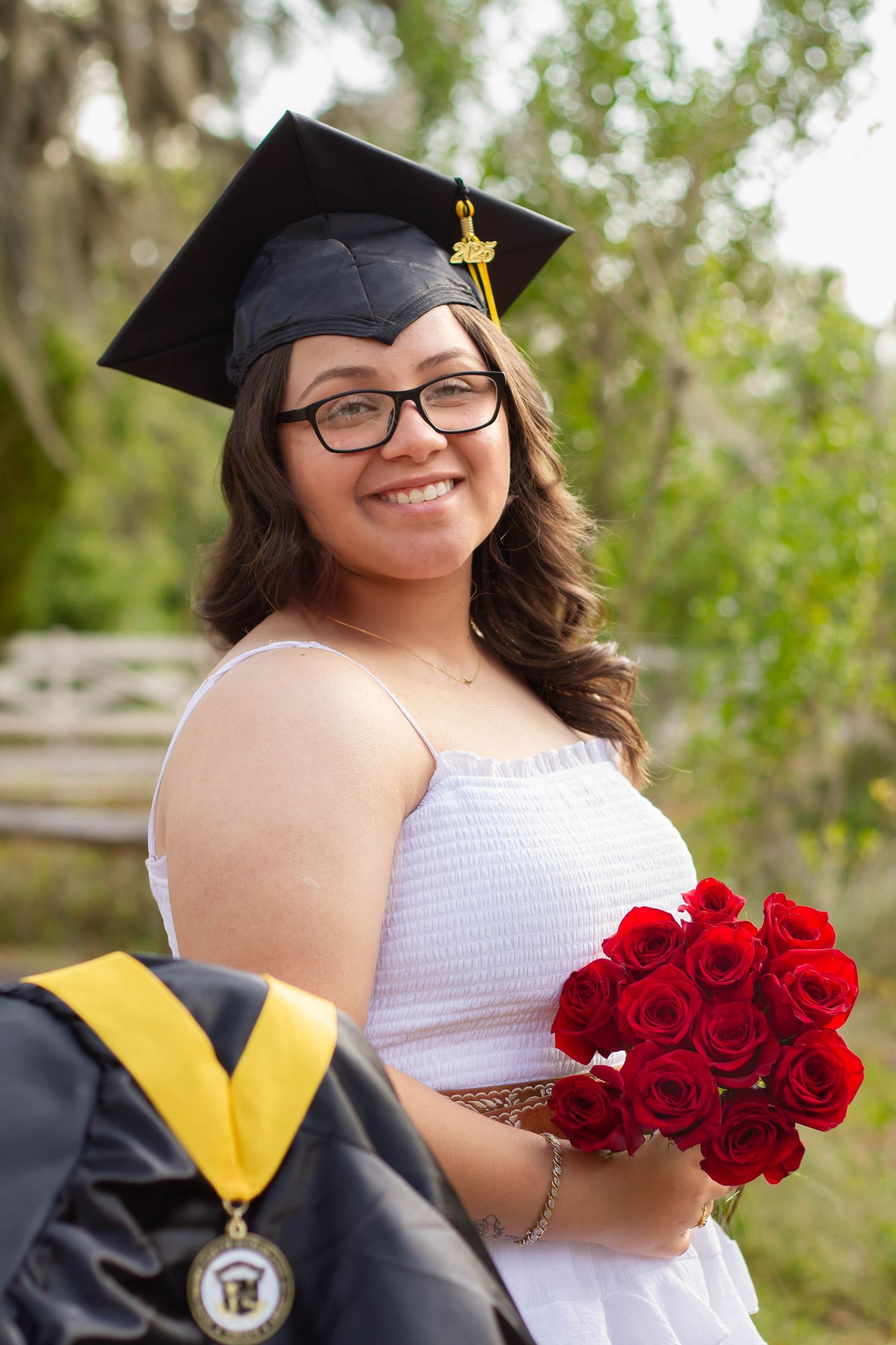 Smiling graduate in black cap holding red roses bouquet wearing white dress outdoors.